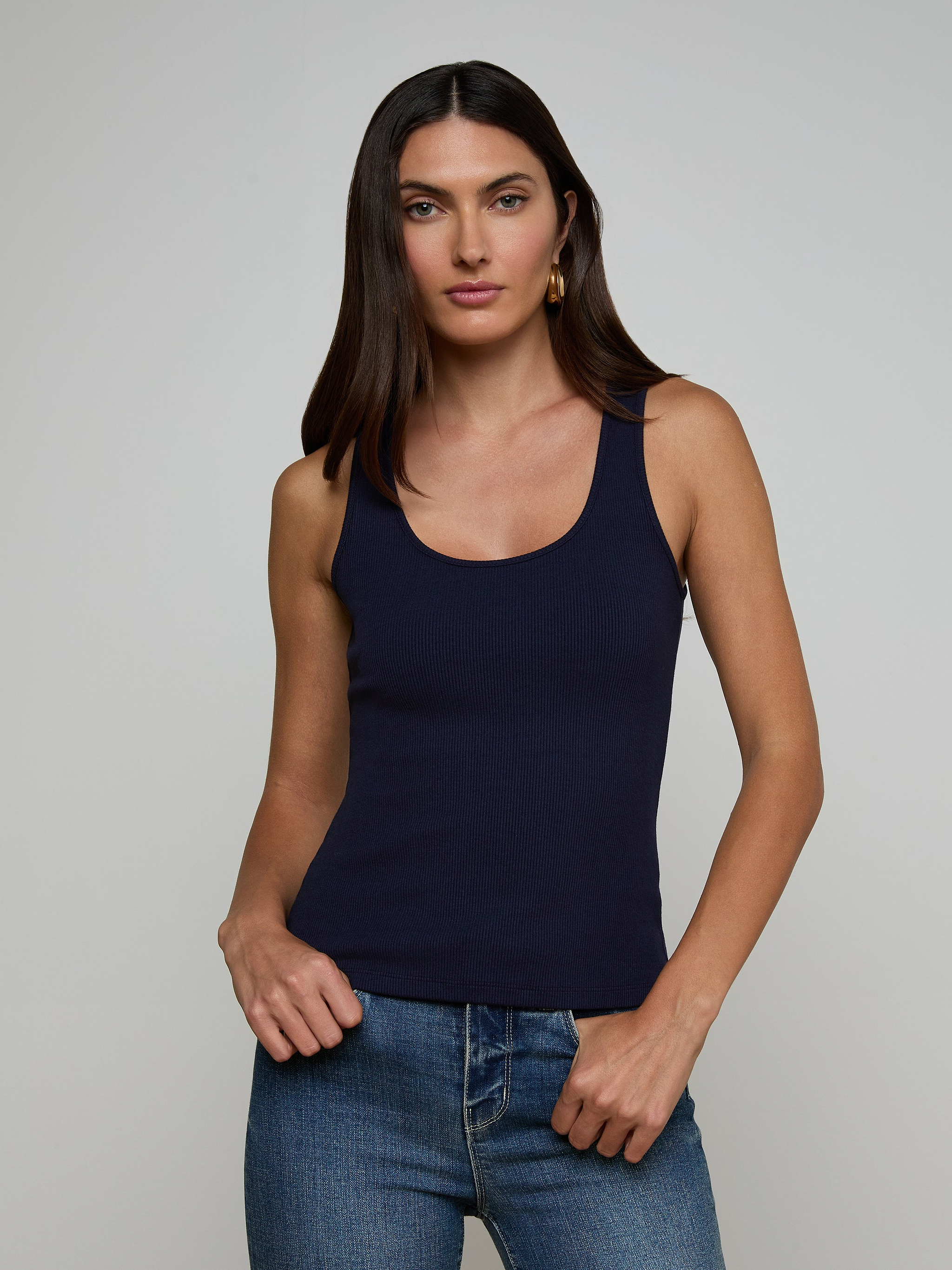 A woman with long dark hair wears the Adira Scoopneck Tank in navy blue and blue jeans, standing against a plain light background, looking at the camera with a neutral expression.