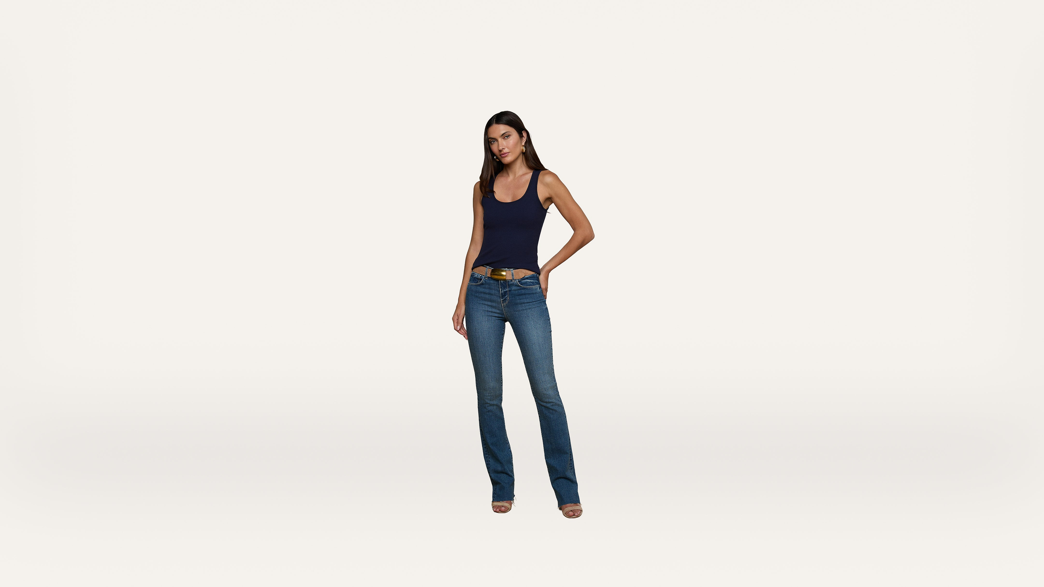 A woman stands facing forward, wearing a navy tank top, blue jeans, and heeled shoes against a plain white background.