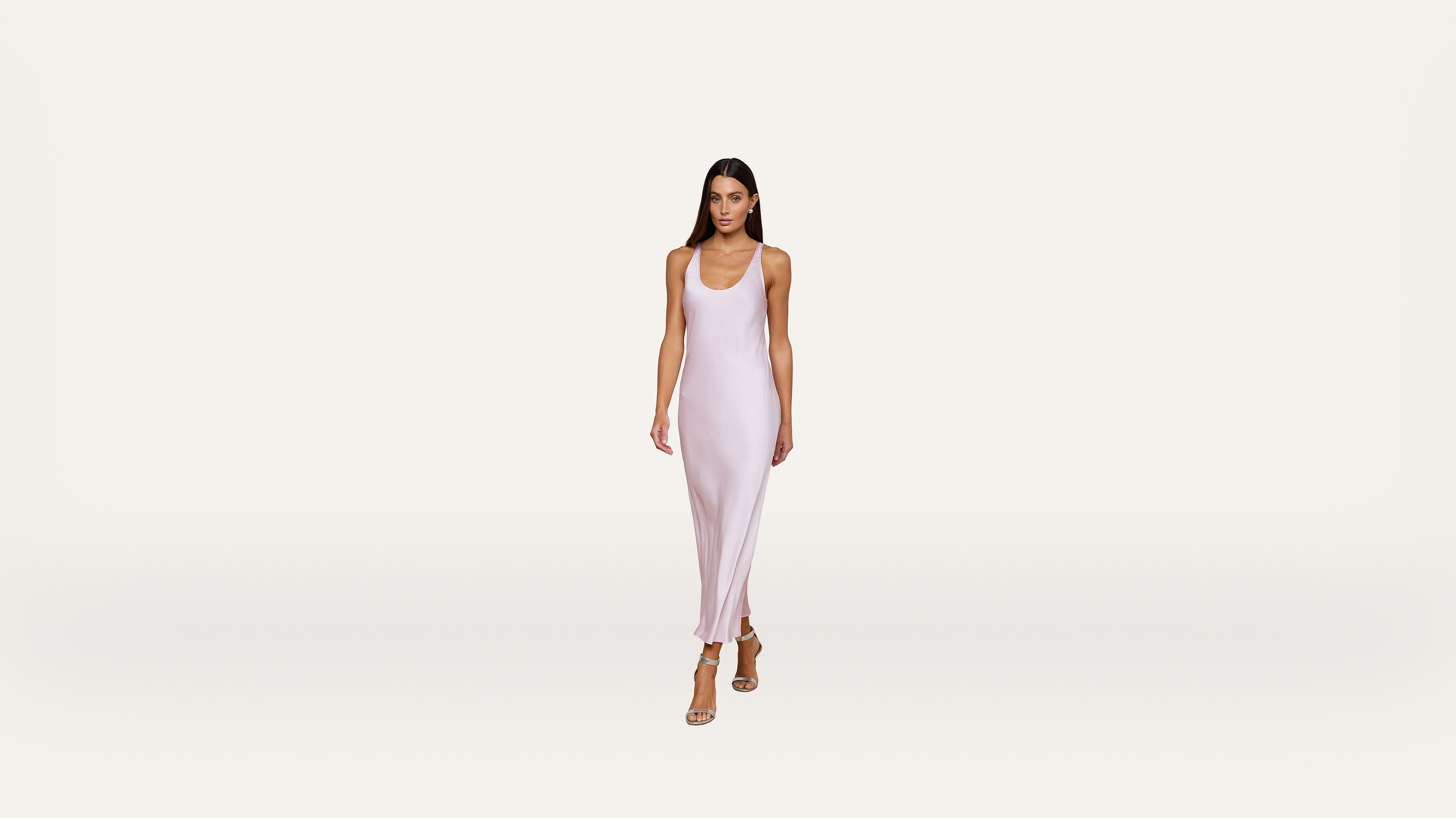 A woman wearing a light pink, sleeveless, ankle-length dress and beige heels walks forward against a plain white background.