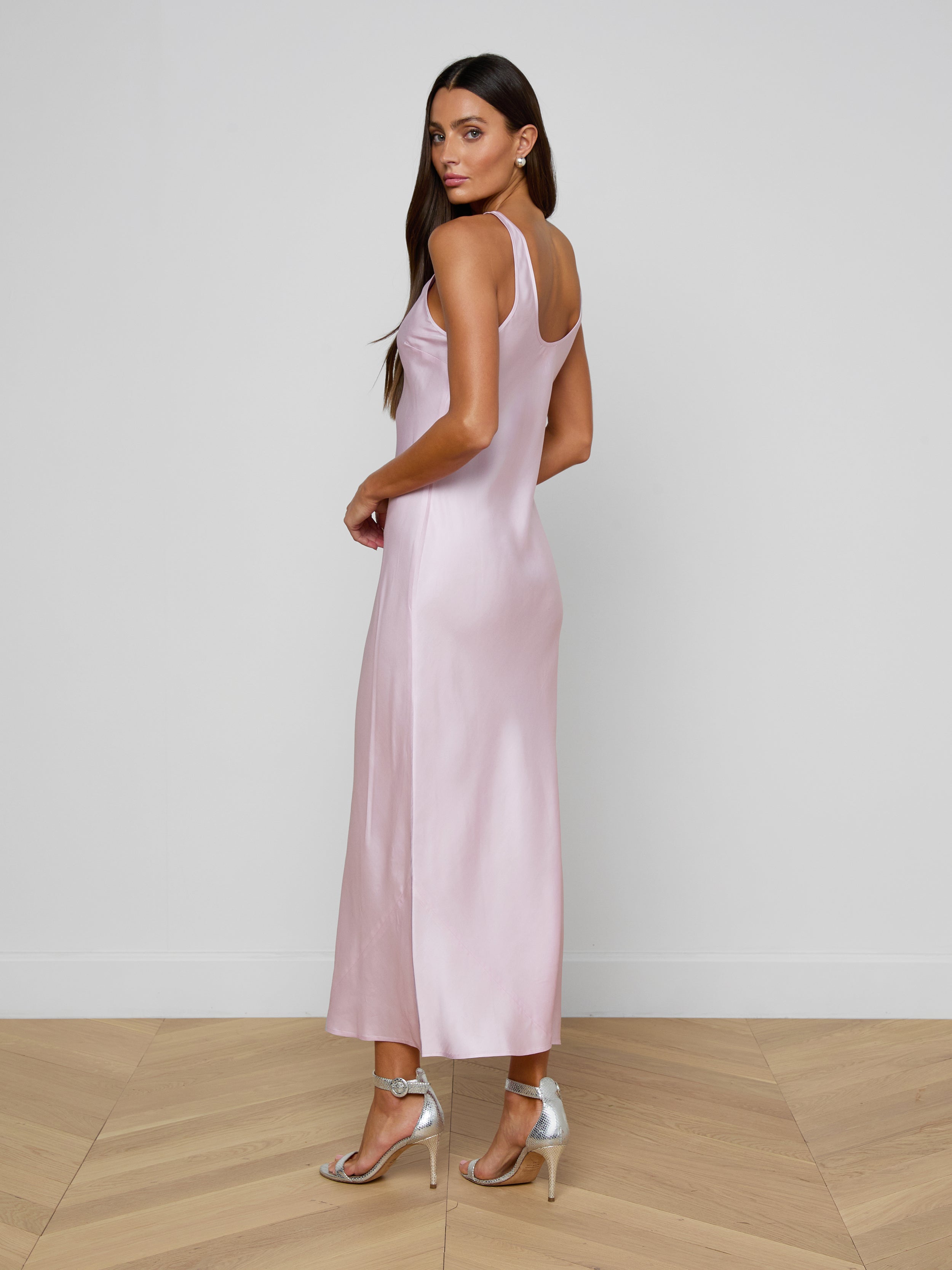 A woman in the Akiya Satin Maxi Dress, featuring a long, bias-cut silhouette in pastel lilac, stands on a plain white background in silver high heels, facing slightly to the side and looking toward the camera.