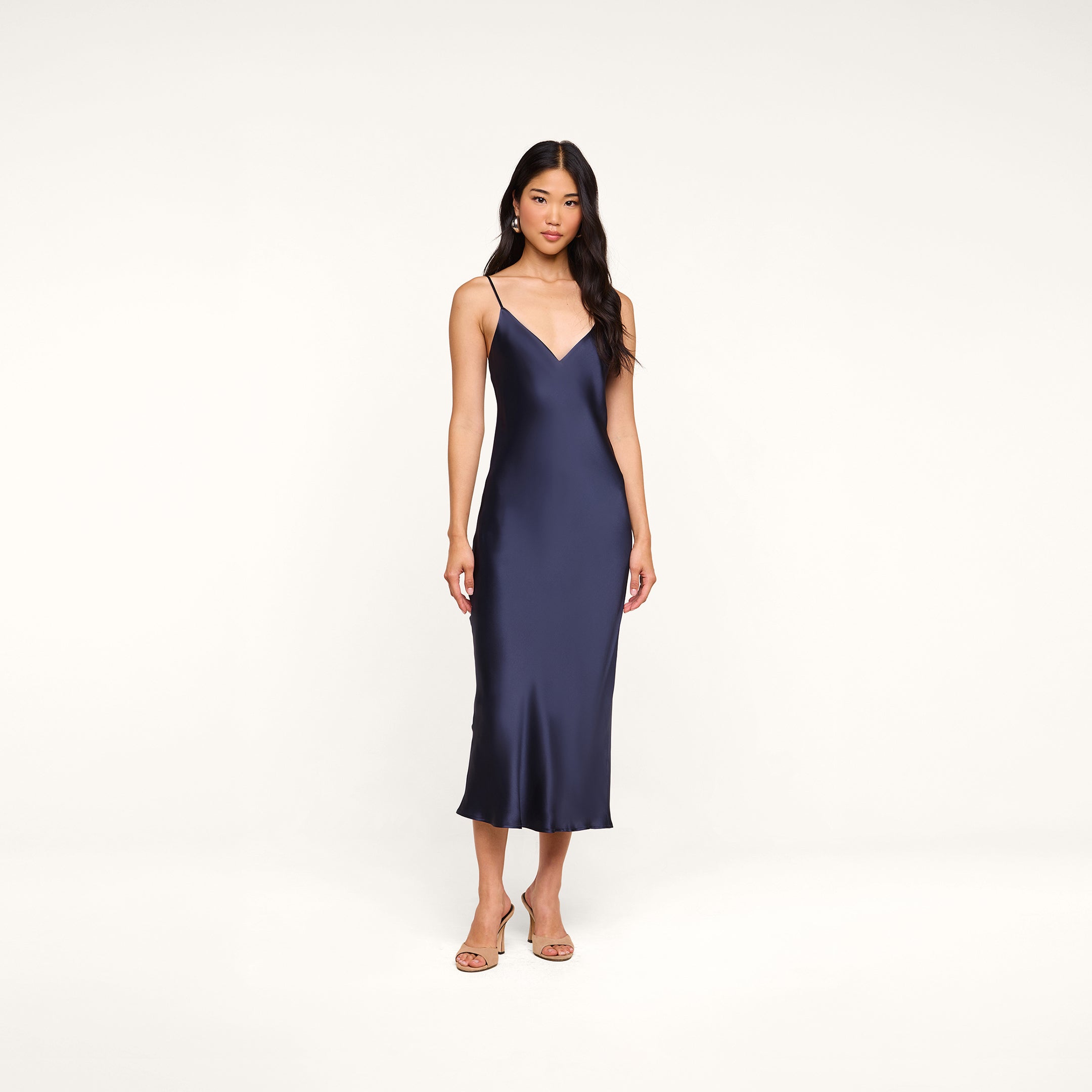 A woman stands against a plain white background wearing the Seridie Silk Slip Dress in navy blue with thin straps, paired with beige heeled sandals—an elegant choice for a wedding guest.