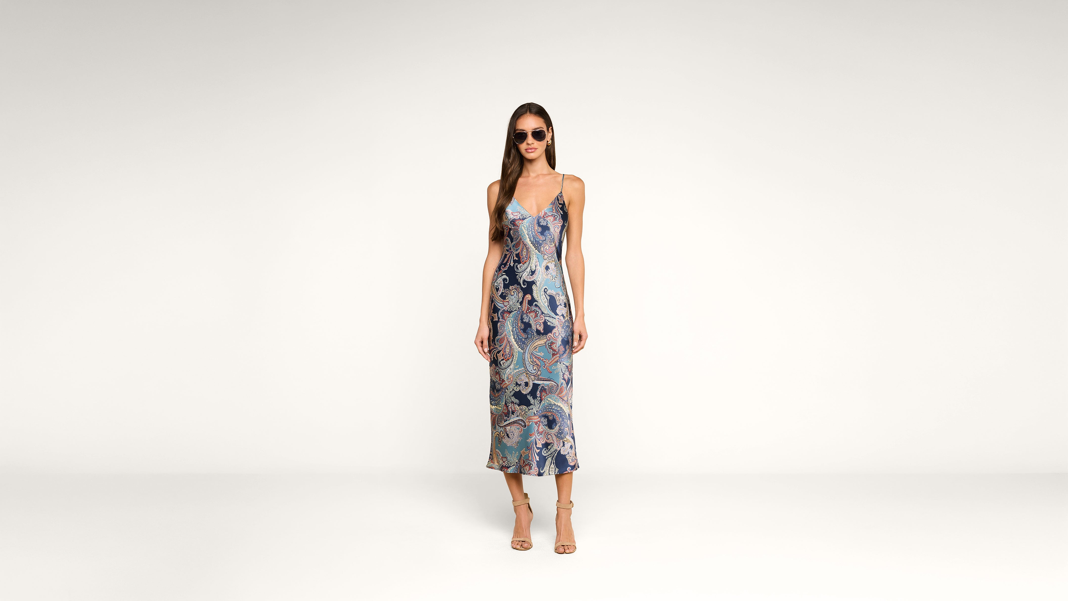 Woman wearing sunglasses and a sleeveless, ankle-length dress with a blue and pink paisley pattern, standing against a plain white background.
