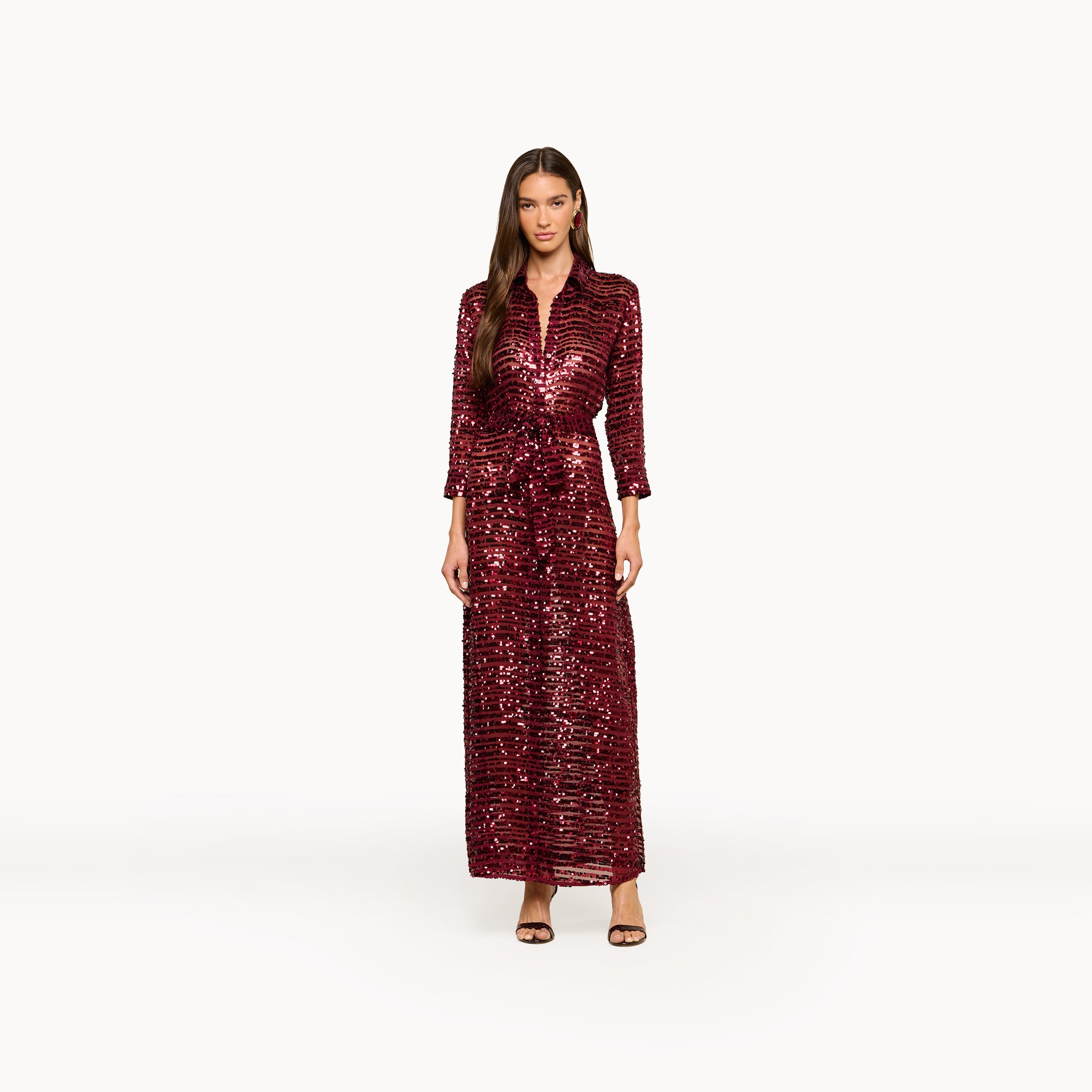 A woman stands against a plain white background wearing the Cameron Sequin Shirt Dress, a maxi-length, deep red sequin dress with long sleeves, paired with black strappy heels.