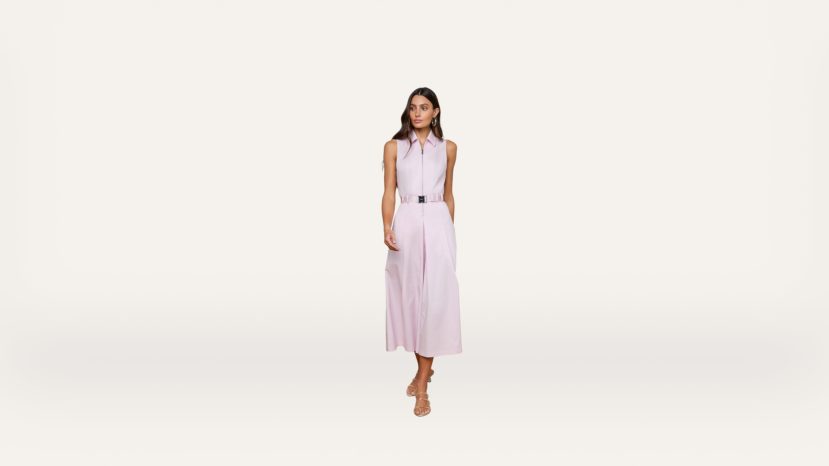 A woman stands against a plain white background, wearing a sleeveless light pink collared midi dress with a belt and beige sandals. She has long brown hair and a neutral expression.