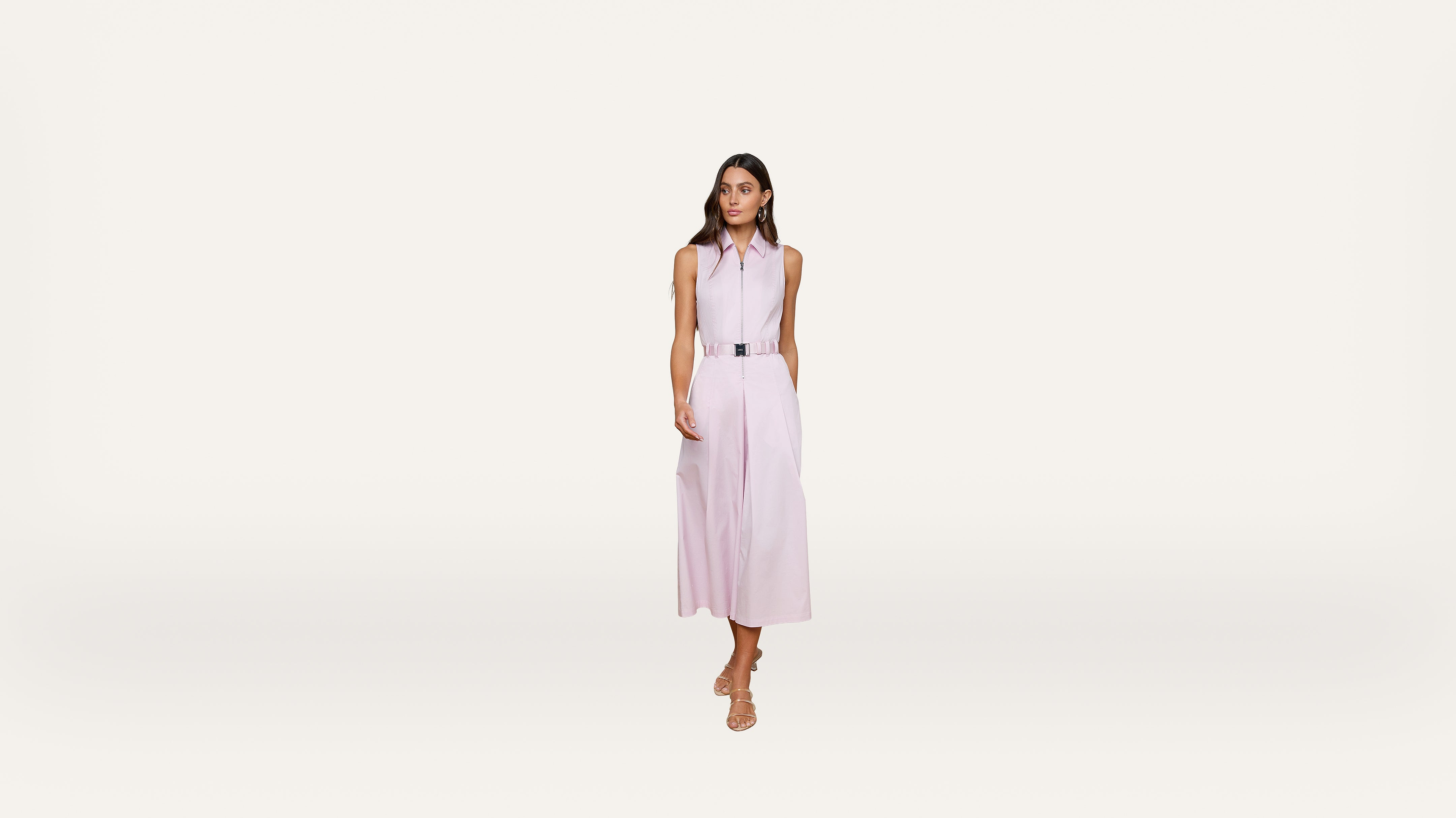 A woman stands against a plain white background, wearing a sleeveless light pink collared midi dress with a belt and beige sandals. She has long brown hair and a neutral expression.