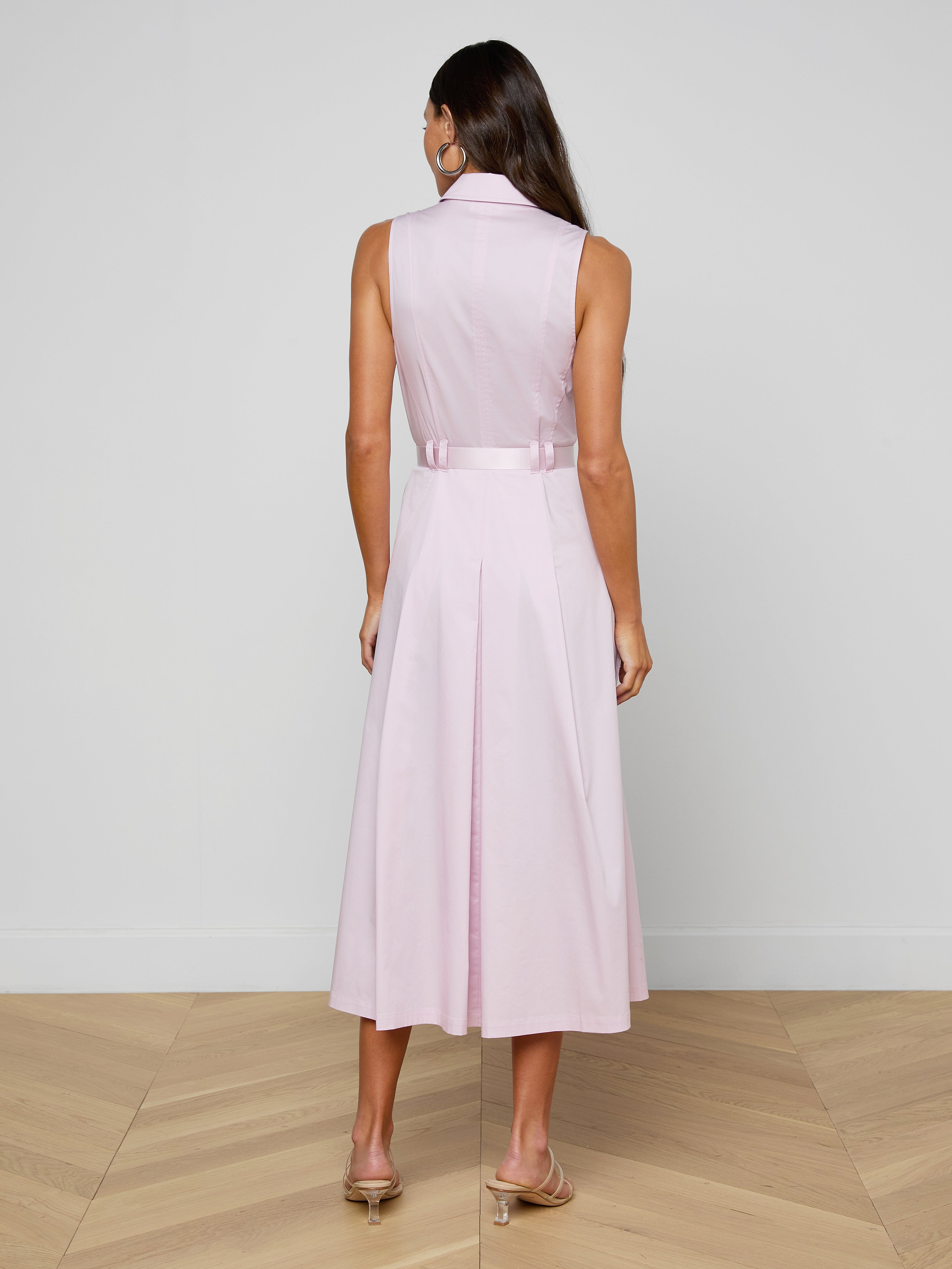 A woman stands with her back to the camera, modeling the sleeveless Kolton Dress in pastel lilac, accented with a statement belt and paired with beige heels against a plain white backdrop.