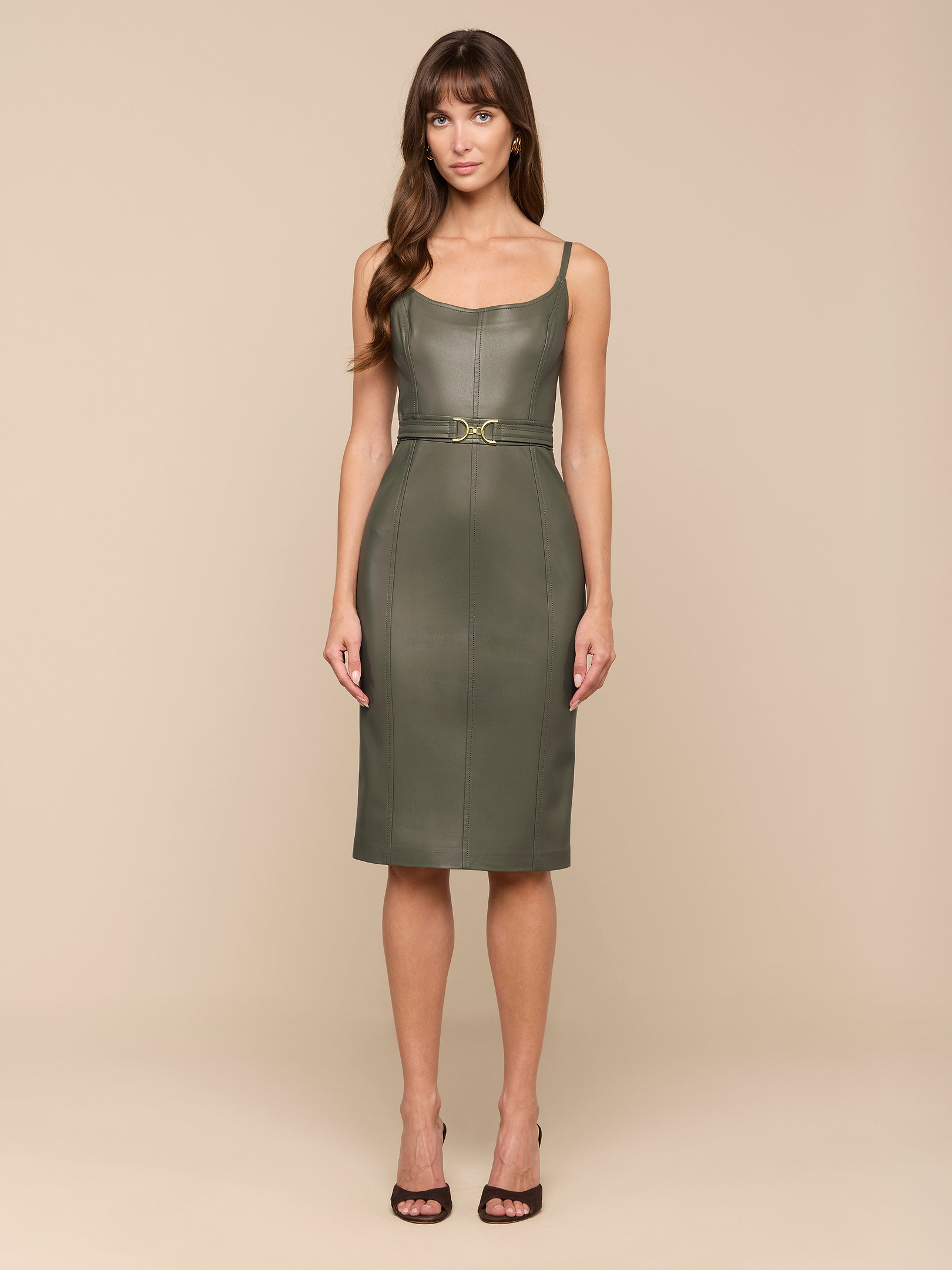 تقف امرأة على خلفية باللون البيج، ووجهها إلى الأمام، وهي ترتدي فستان Halia Vegan Leather Dress باللون الزيتوني الدخاني - وهو فستان متوسط الطول بلا أكمام مع حزام خصر - مع صندل أسود بكعب عالٍ.