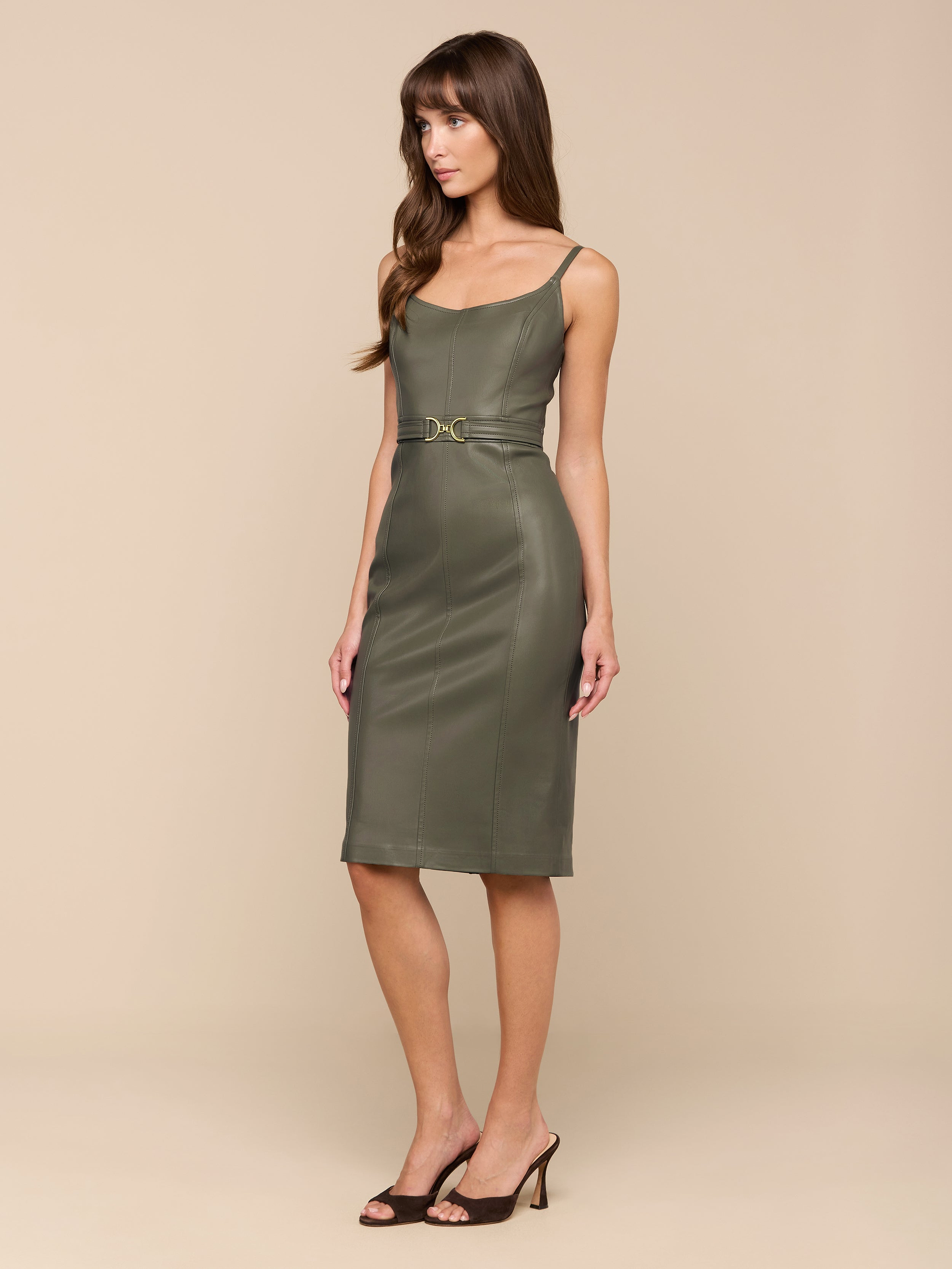 امرأة تقف على خلفية باللون البيج وهي ترتدي فستان Halia الجلدي النباتي Halia باللون الزيتوني الدخاني. يتميز هذا الفستان متوسط الطول بحمالات رفيعة وحزام خصر. لديها شعر بني طويل وتنسق الإطلالة مع حذاء أسود مفتوح من الأمام.