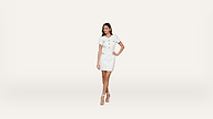 A woman stands confidently in a short white dress with gold buttons and white high-heeled sandals against a plain, light background.