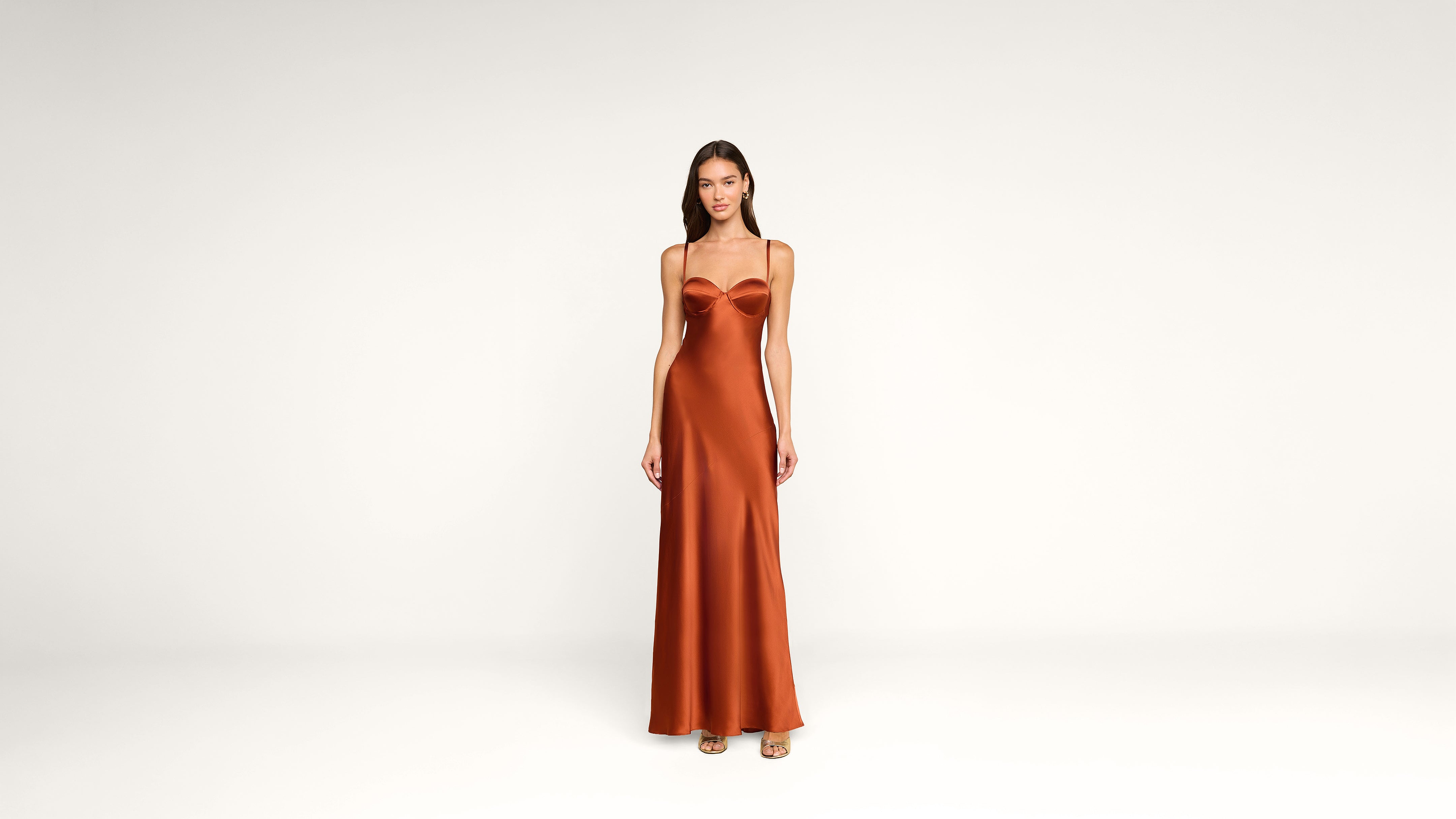A woman stands facing forward, wearing a long, copper-colored satin dress with spaghetti straps against a plain white background.