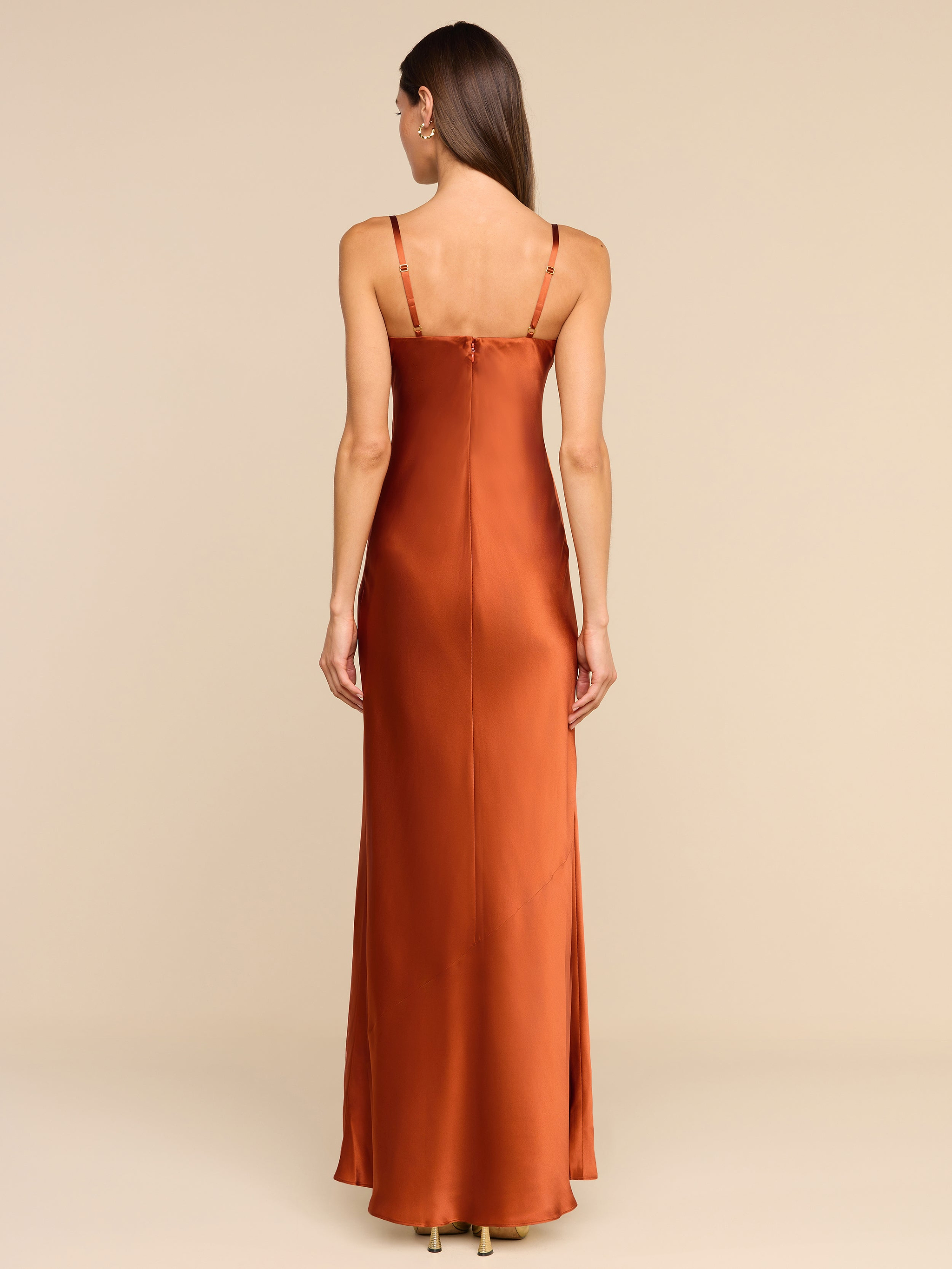 A woman stands facing away, wearing the Sayla Silk Gown—a long, copper silk dress with thin straps. The plain white background and her straight, loose hair cascading down her back create an event-ready look infused with vintage glamour.