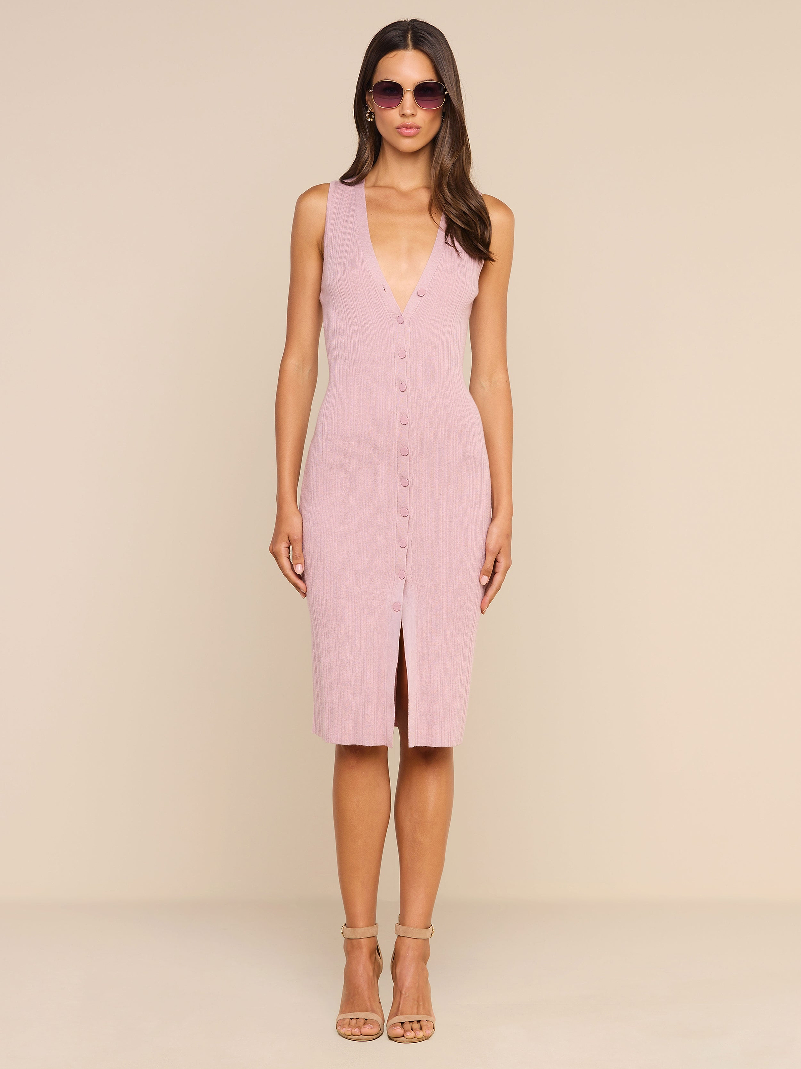Una donna è in posa su uno sfondo beige a tinta unita e indossa il Domino Dress — un abito midi senza maniche di colore rosa chiaro, con chiusura frontale a bottoni, scollo a V e spacco sul davanti — abbinato a tacchi color carne e occhiali da sole rotondi.