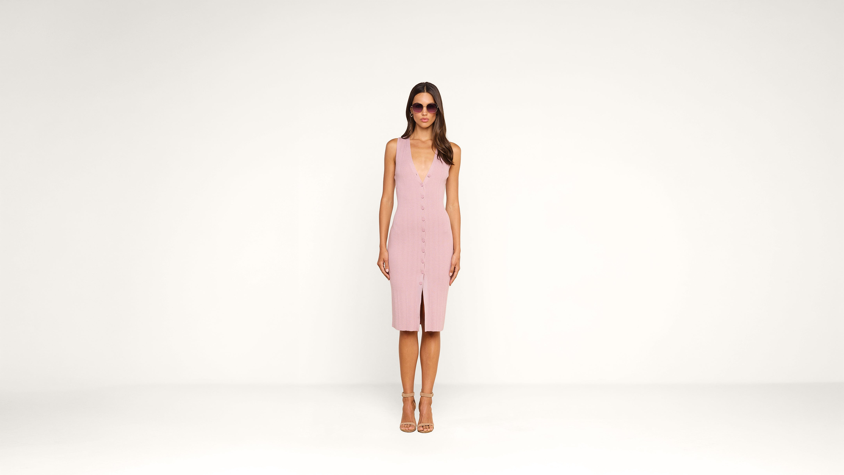 A woman wearing sunglasses and a sleeveless pink dress with a front slit stands facing forward against a plain white background.