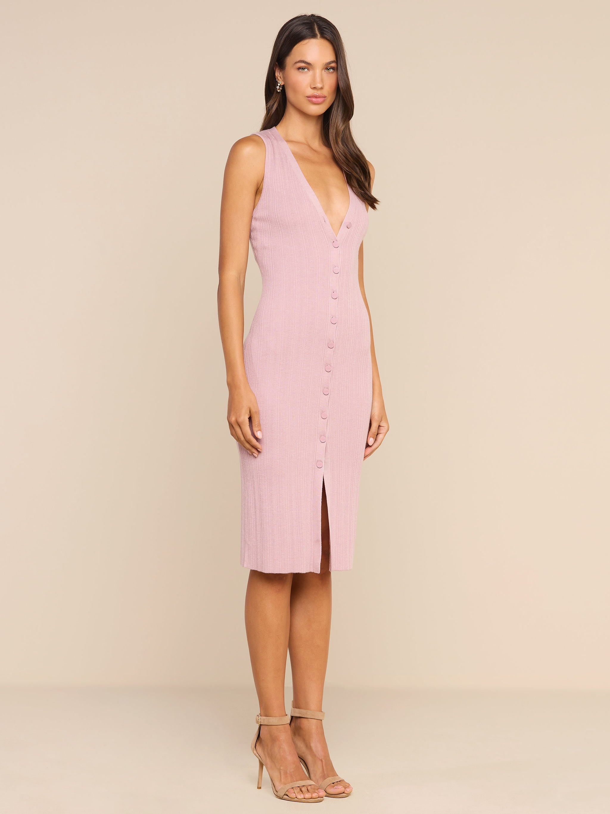 A woman stands in a studio wearing the Domino Dress—a sleeveless, light pink midi dress featuring a button-down front, V-neckline, and front slit. She has long hair and pairs the look with strappy high-heeled sandals.