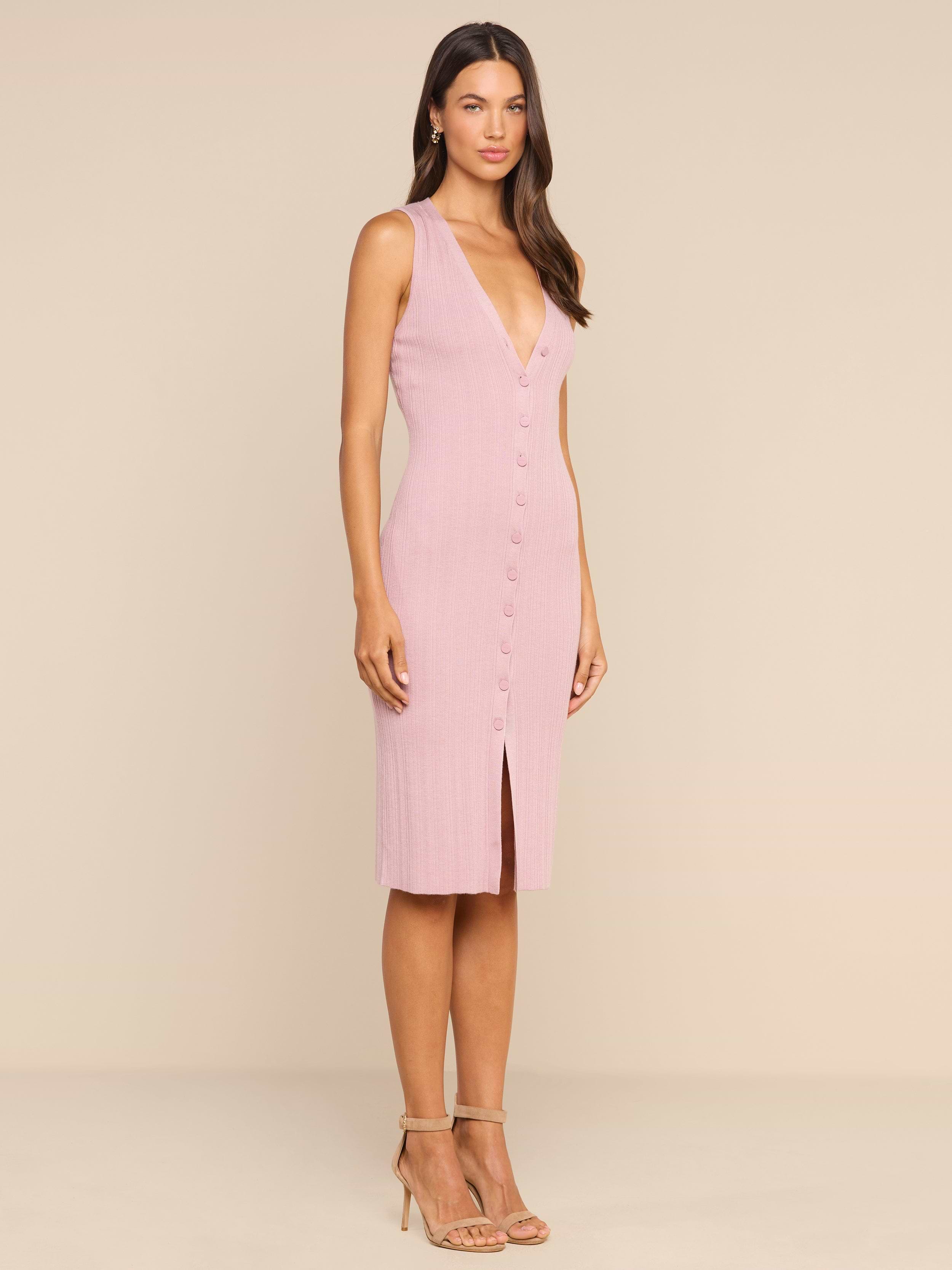 A woman stands in a studio wearing the Domino Dress—a sleeveless, light pink midi dress featuring a button-down front, V-neckline, and front slit. She has long hair and pairs the look with strappy high-heeled sandals.