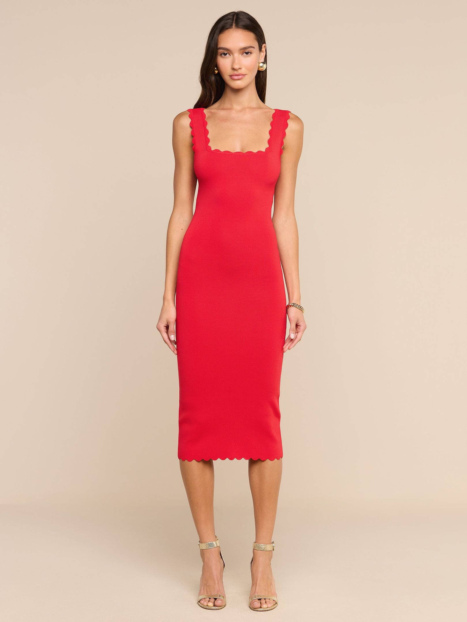 Una donna è in posa su uno sfondo beige e indossa l’abito in maglia Romona: un miniabito senza maniche, dalla linea aderente, di colore rosso rossetto, caratterizzato da scollo quadrato smerlato e orlo smerlato. Ha i capelli lunghi e abbina il look a sandali beige con tacco alto e cinturini e a un braccialetto.