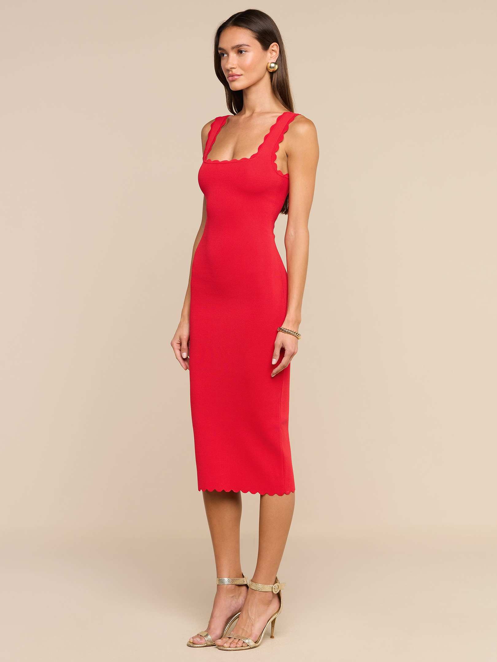 Donna che indossa un abito in maglia Romona senza maniche e dalla linea aderente, di colore rosso rossetto con bordi smerlati, abbinato a orecchini a cerchio dorati, braccialetti e sandali dorati con tacco alto, in piedi su uno sfondo beige.