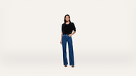 A woman stands facing forward, wearing a black top, blue high-waisted wide-leg jeans, and black heeled sandals, against a plain white background.