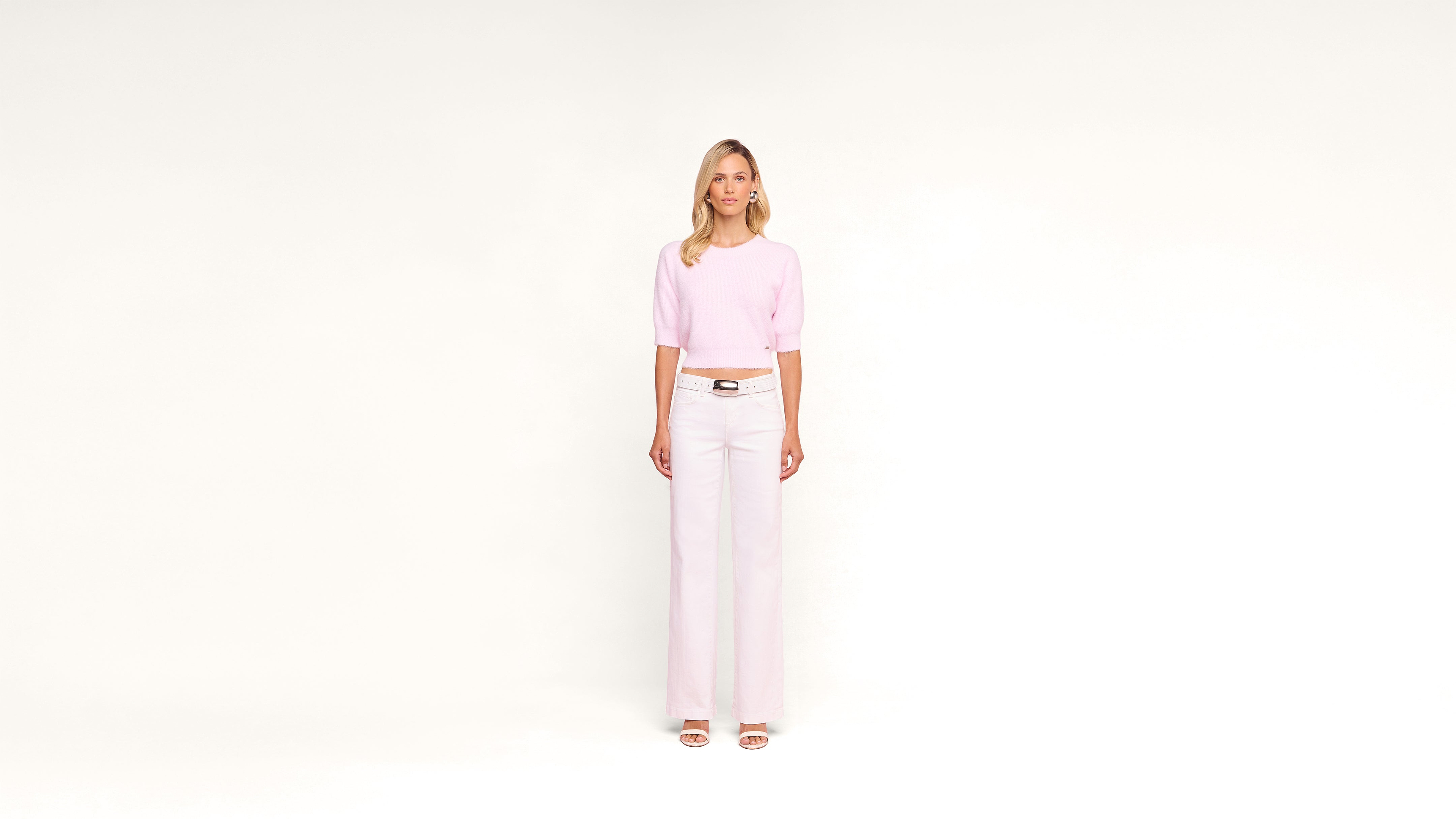 A woman stands against a plain white background, wearing a light pink top, white high-waisted wide-leg pants, and white sandals. She faces forward with a neutral expression.