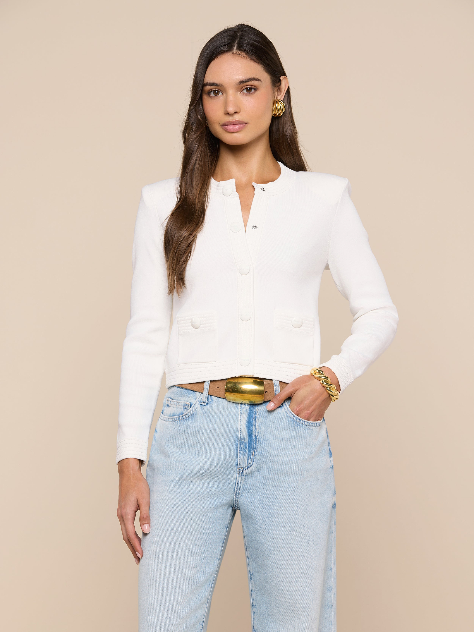 A woman with long brown hair wears the Patsy Cropped Cardigan—a white, buttoned style with subtle shoulder pads—paired with light blue jeans and gold accessories. She stands facing forward against a plain light background with a neutral expression.