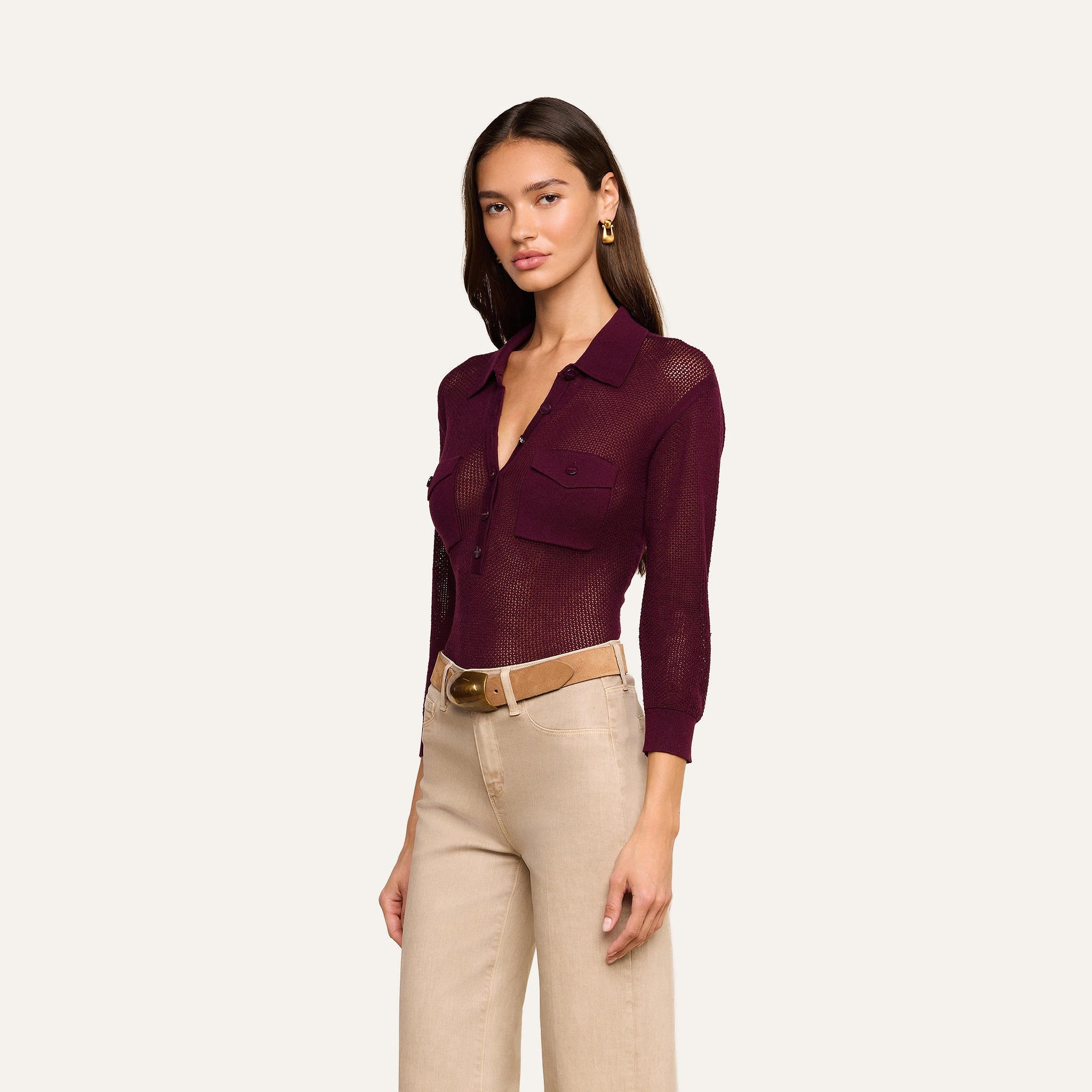Wearing the Carine Top, a sheer burgundy button-up with utility pockets, paired with beige high-waisted pants, a tan belt, and gold earrings, she stands against a plain background.