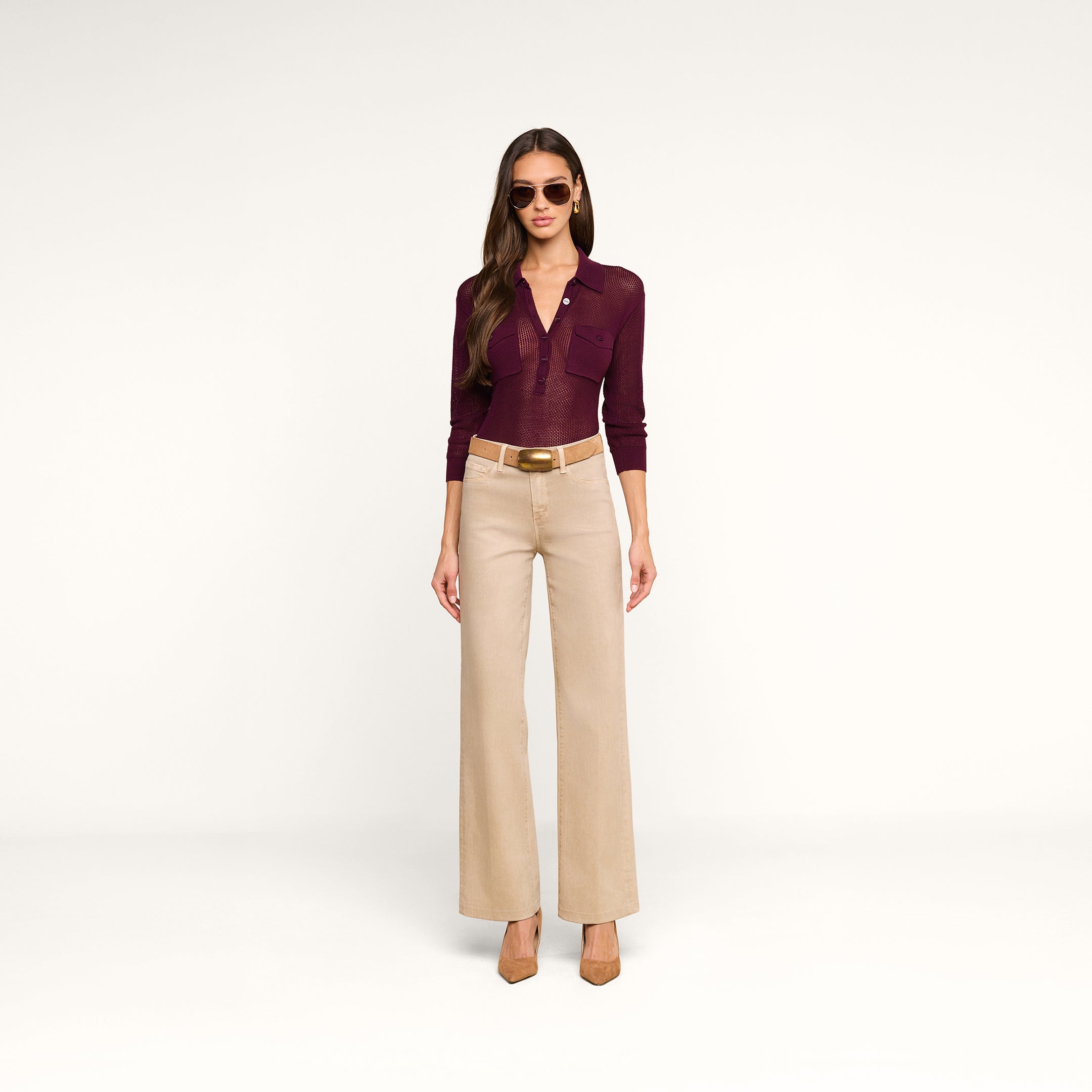 A woman wearing sunglasses, a burgundy Carine Top, beige wide-leg pants with utility pockets, a white belt, and brown heels stands against a plain white background.