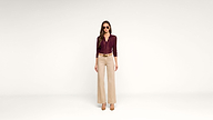 A woman wearing sunglasses, a maroon button-up top, beige high-waisted wide-leg pants, and brown heels stands against a plain white background.