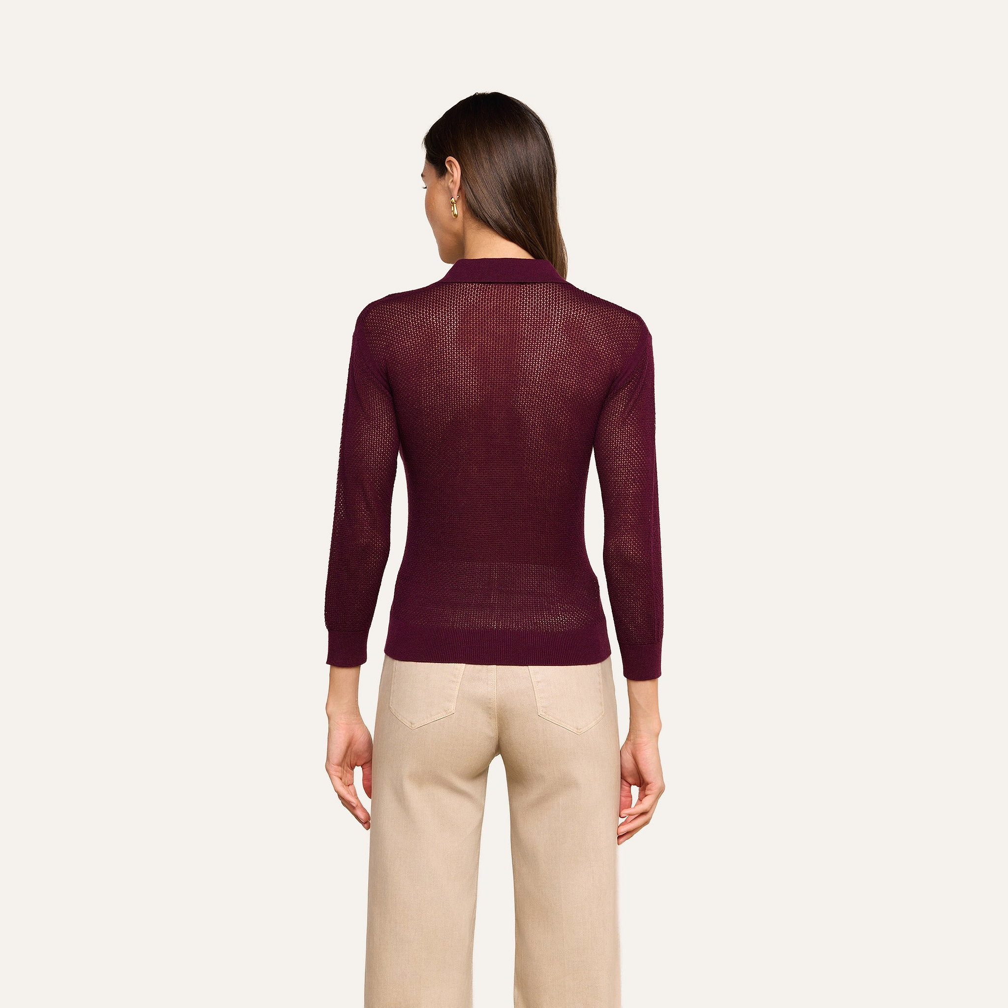 A woman stands facing away, wearing a lightweight knit maroon Carine Top with long sleeves and beige pants. Her straight brown hair is down, and her arms hang at her sides against a plain light background.