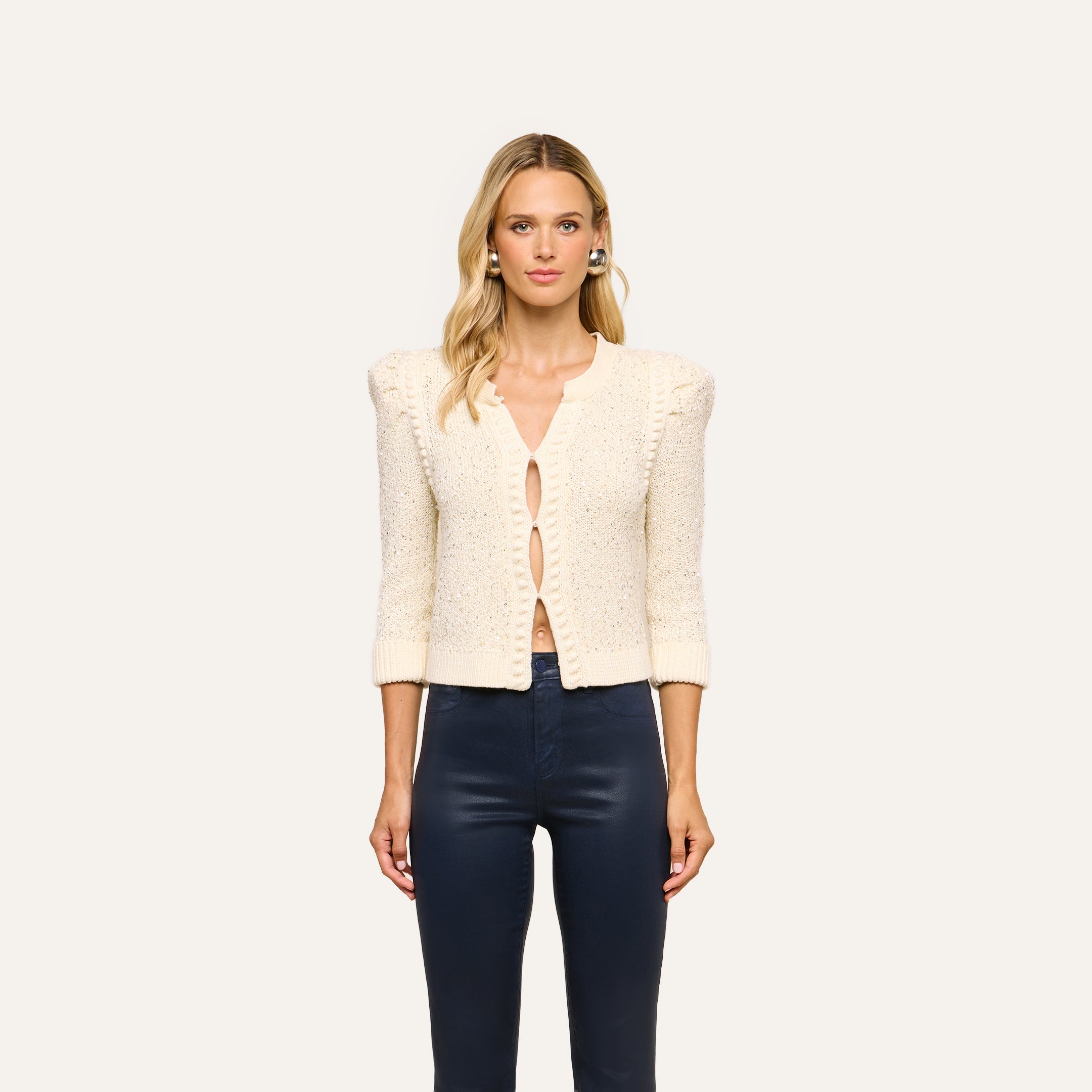A woman stands against a plain light background, facing forward with a neutral expression, wearing the Thistle Sequined Cardigan in ivory—collarless and embellished with sequins—paired with large pearl earrings and navy high-waisted pants.