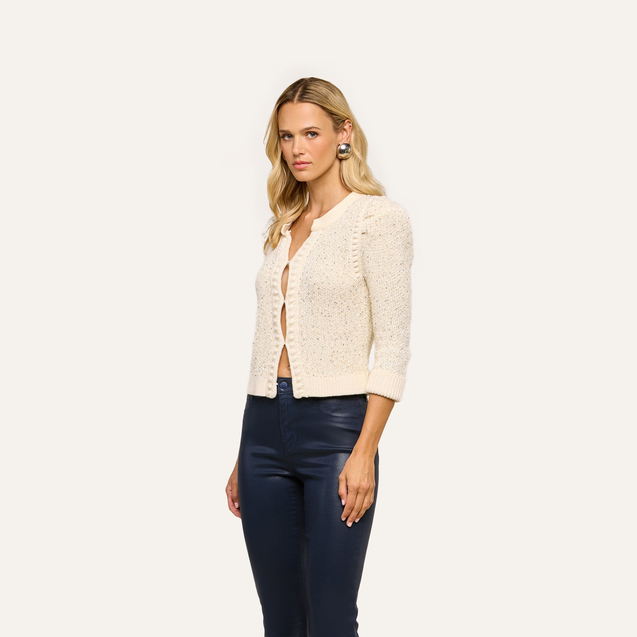A woman with blonde hair wears the Thistle Sequined Cardigan in ivory, styled over a beige top and navy blue pants, standing against a plain light background.