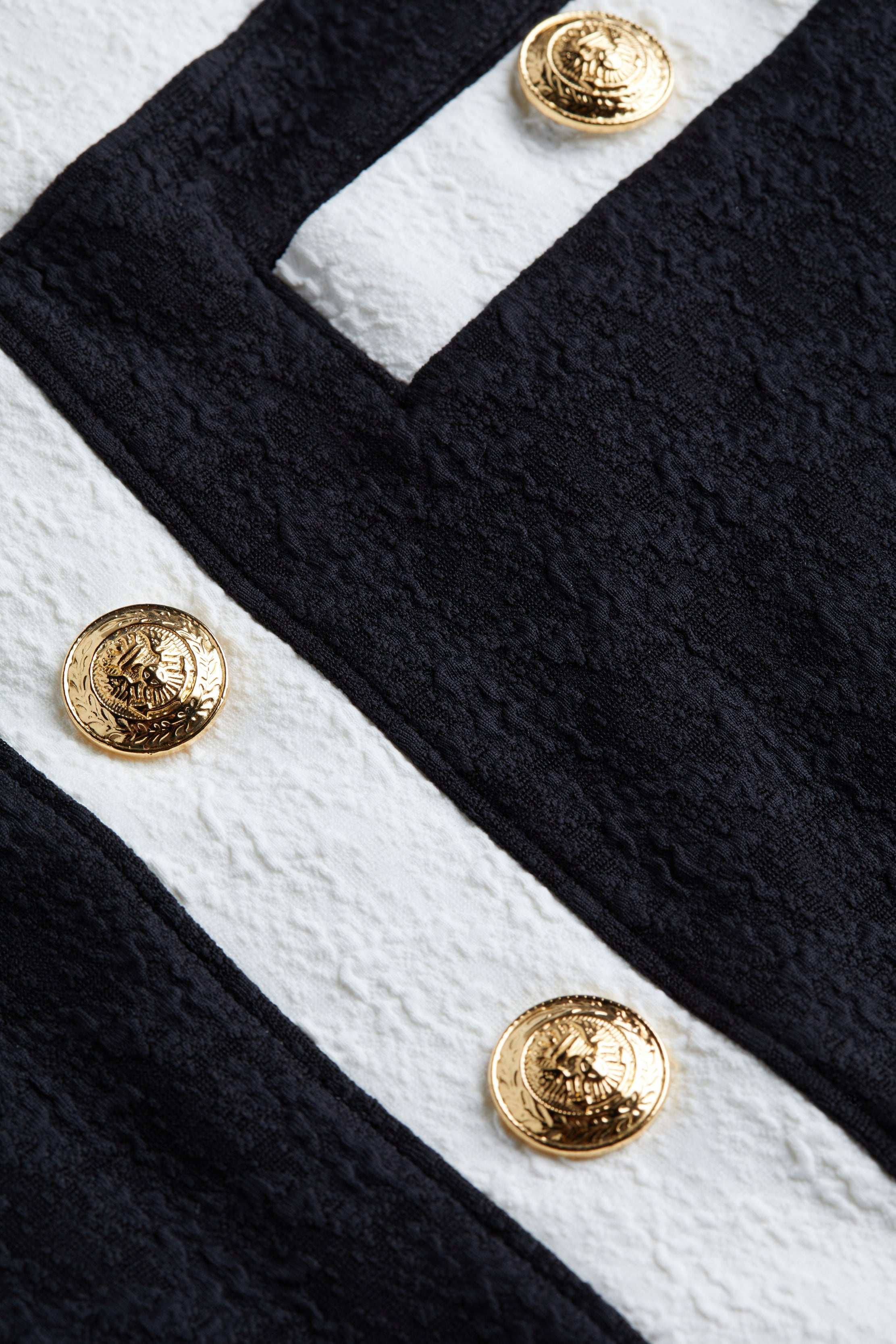 Close-up of the Coco Mini Skirt Cover-Up’s textured bouclé fabric in black and white, accented with gold decorative buttons. The embossed design on the buttons adds a luxurious detail to the garment.