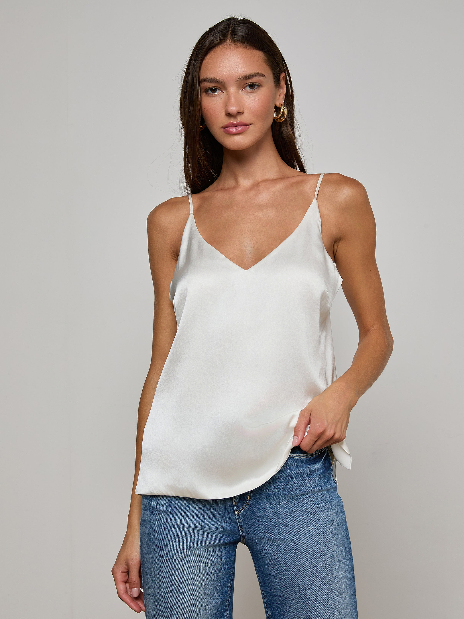 A woman wearing the Gabriella Silk Camisole in ivory and blue jeans stands against a plain light background, looking at the camera with a neutral expression.