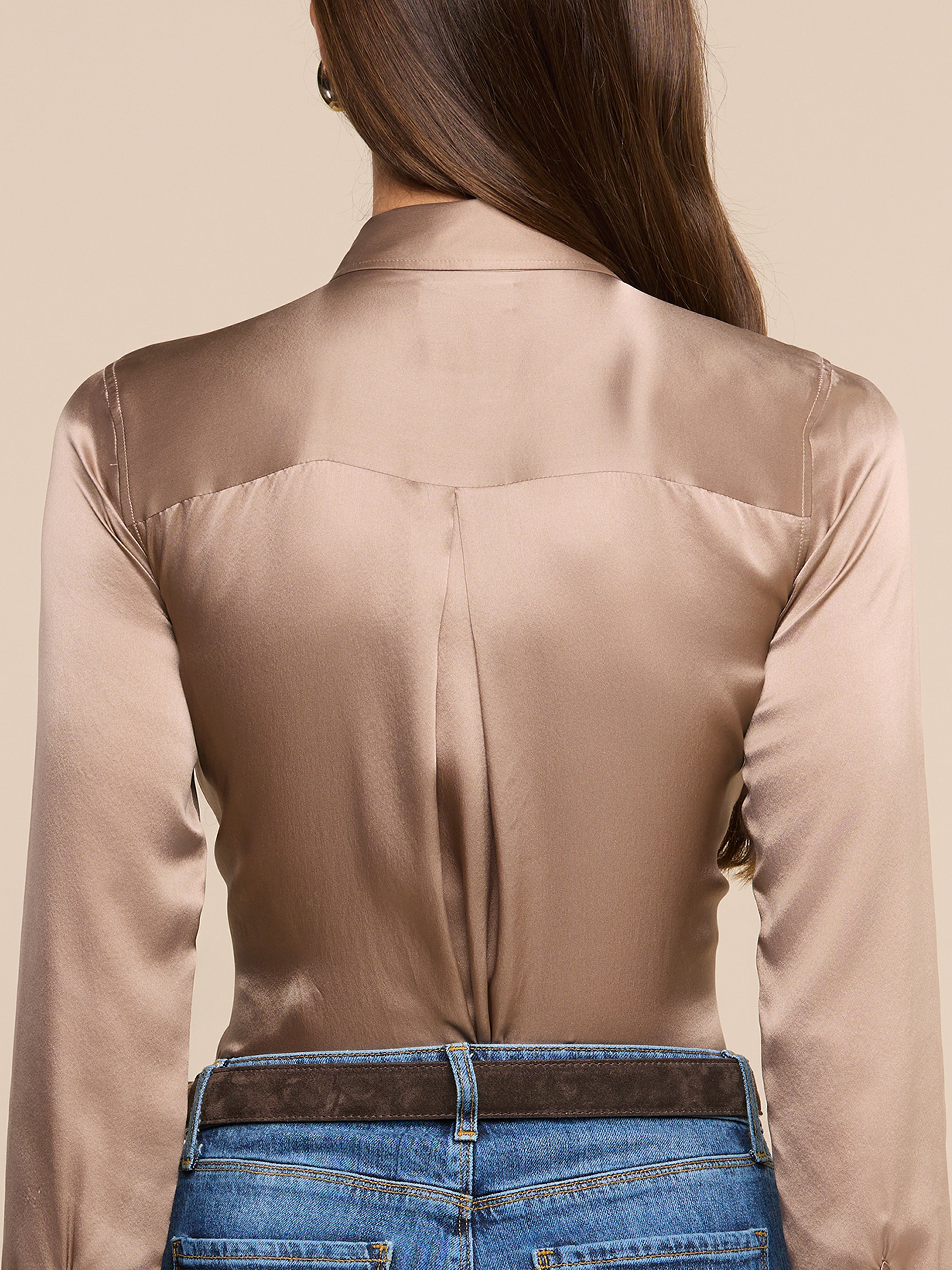A woman with long brown hair wears a shiny light brown blouse with long sleeves and blue jeans. The image shows her from the back, highlighting the blouses smooth texture and tailored fit.