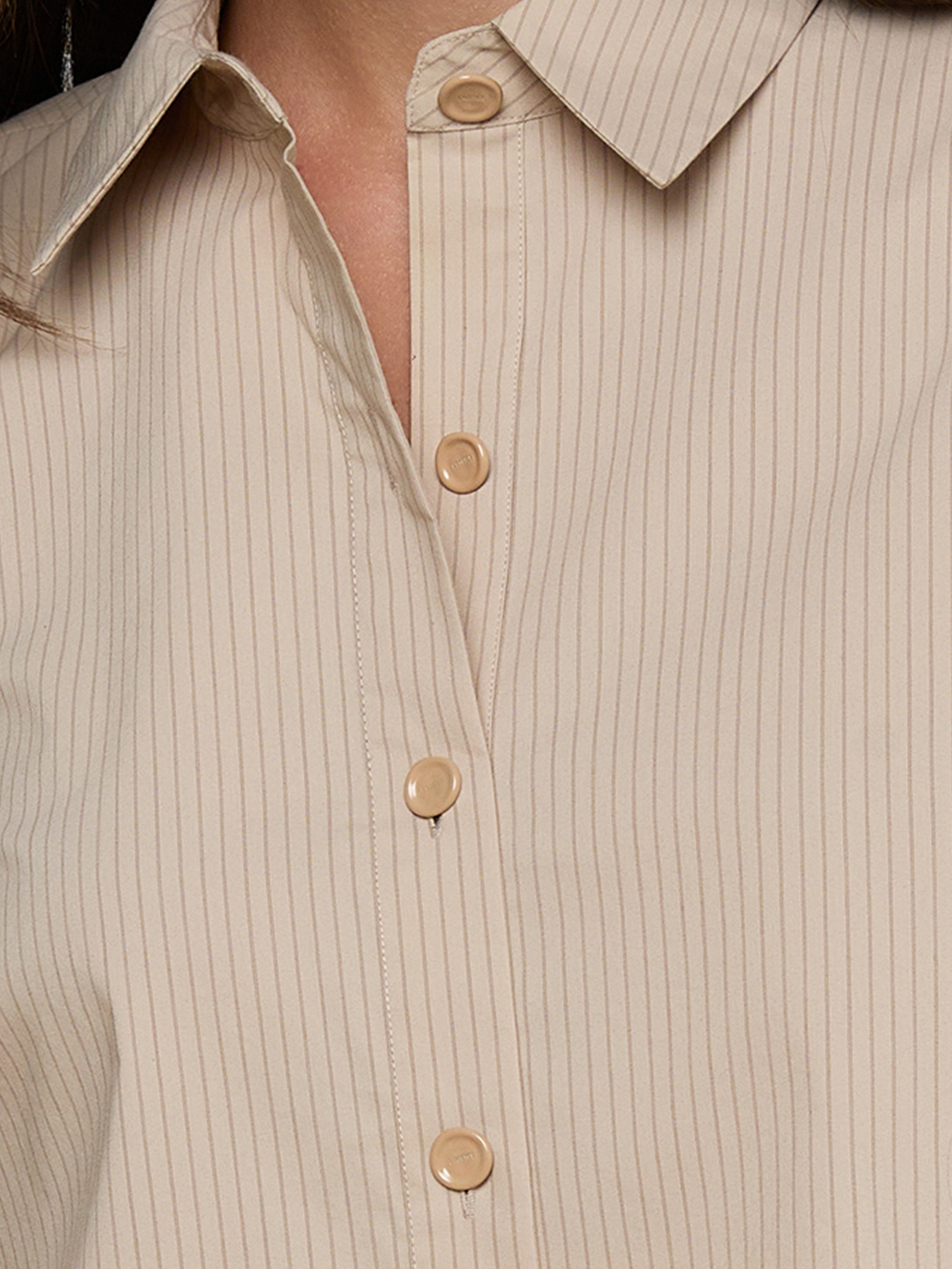 Close-up of a person wearing a beige, collared shirt with thin vertical stripes and light brown buttons. The shirt is partially buttoned up. The persons lower face and neck are slightly visible.