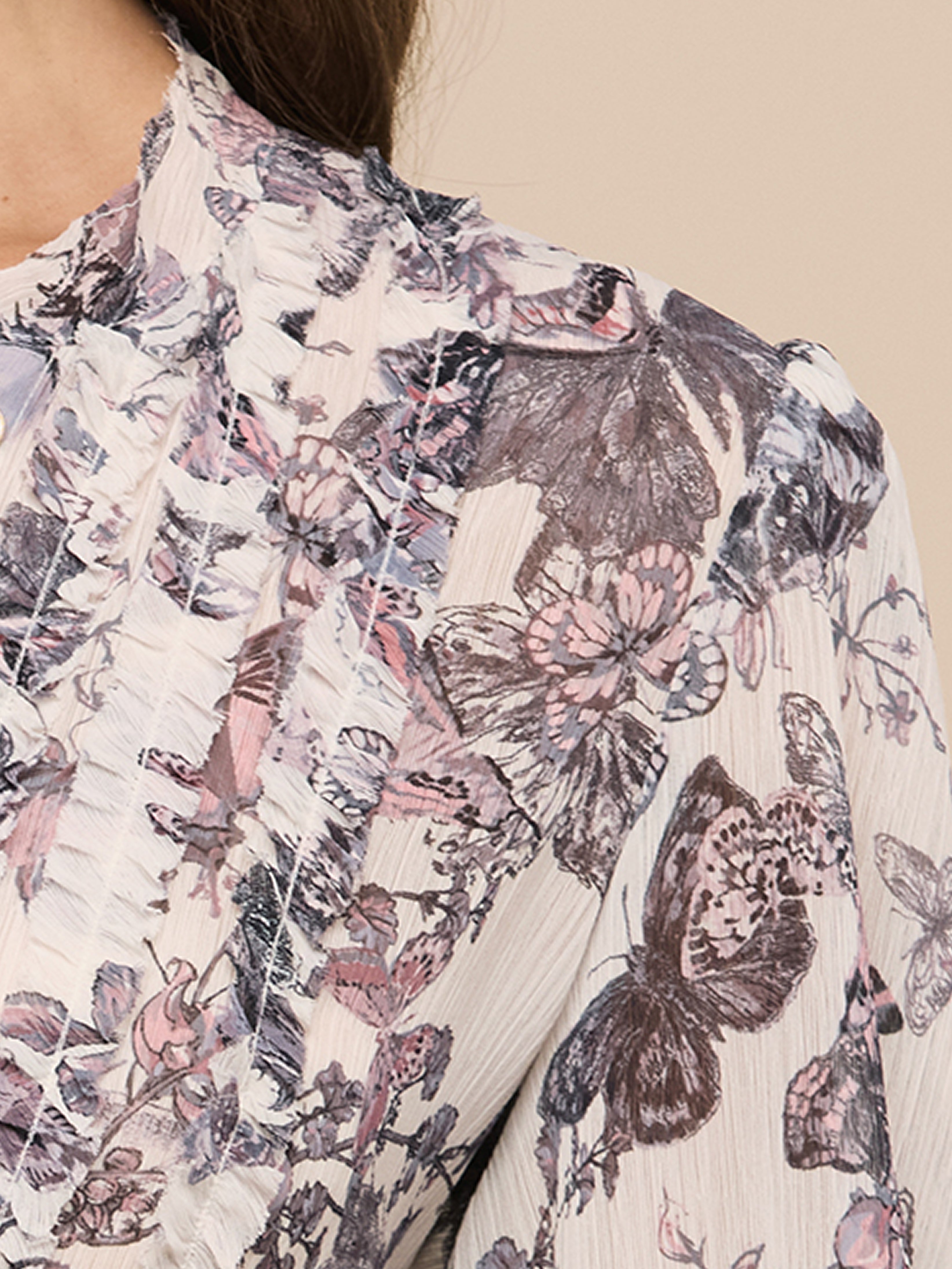 Close-up of a floral patterned blouse with ruffled detailing. The fabric features pink, purple, and gray flowers and butterflies on a light background. Only part of the wearer’s shoulder, neck, and hair is visible.