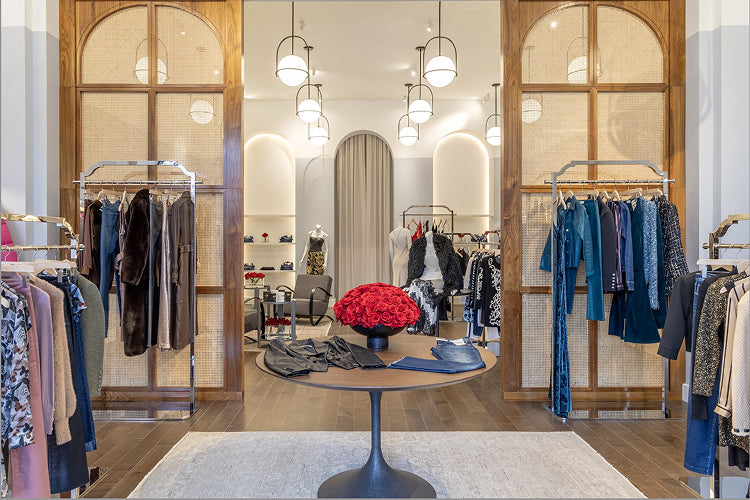 A stylish clothing boutique with wooden floors, elegant lighting, racks of assorted women’s fashion, and a round table in the center topped with a bouquet of red roses and folded garments.