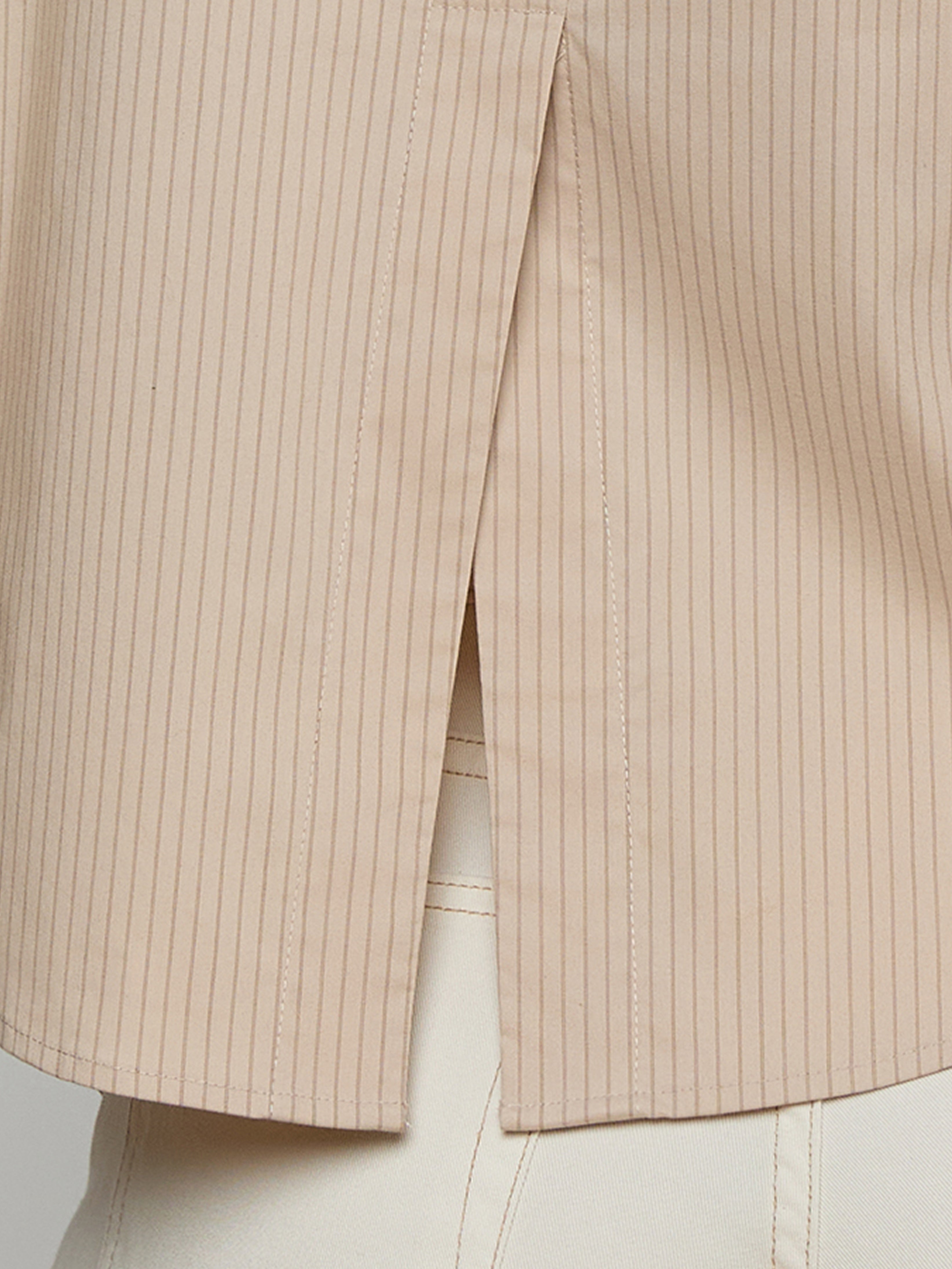 Close-up of a beige pinstriped shirt with a side slit hem, worn over high-waisted cream pants.