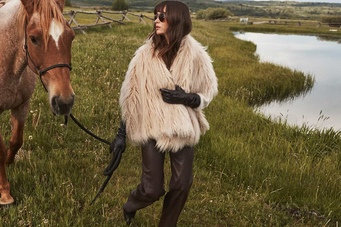 A person wearing a fluffy cream-colored coat, black gloves, and sunglasses stands on grass near a pond, holding the reins of a brown horse.