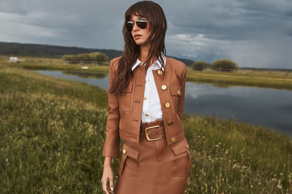 A woman in a tan jacket and skirt stands in a grassy field near a pond, wearing sunglasses and a white shirt. The sky is cloudy, and trees are visible in the background.