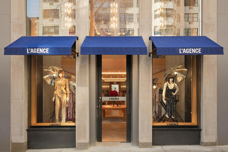 A clothing store front with two blue awnings labeled L’AGENCE. Two mannequins in fashionable outfits are displayed in the windows on either side of the glass door entrance. The store interior is visible inside.