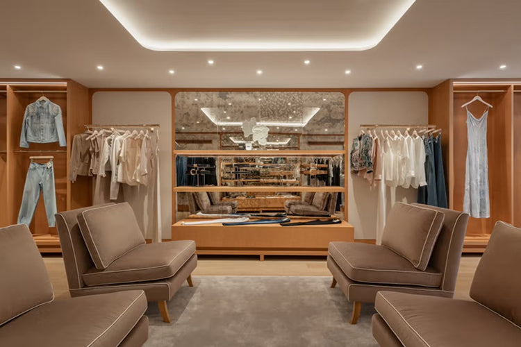 A spacious, modern walk-in closet with beige armchairs, wooden shelves displaying neatly hung clothes, a large mirror, and soft recessed lighting creating an elegant atmosphere.