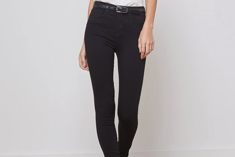 A person stands against a light wall, wearing a white shirt tucked into high-waisted black skinny jeans with a black belt. Only their body from shoulders down is visible.