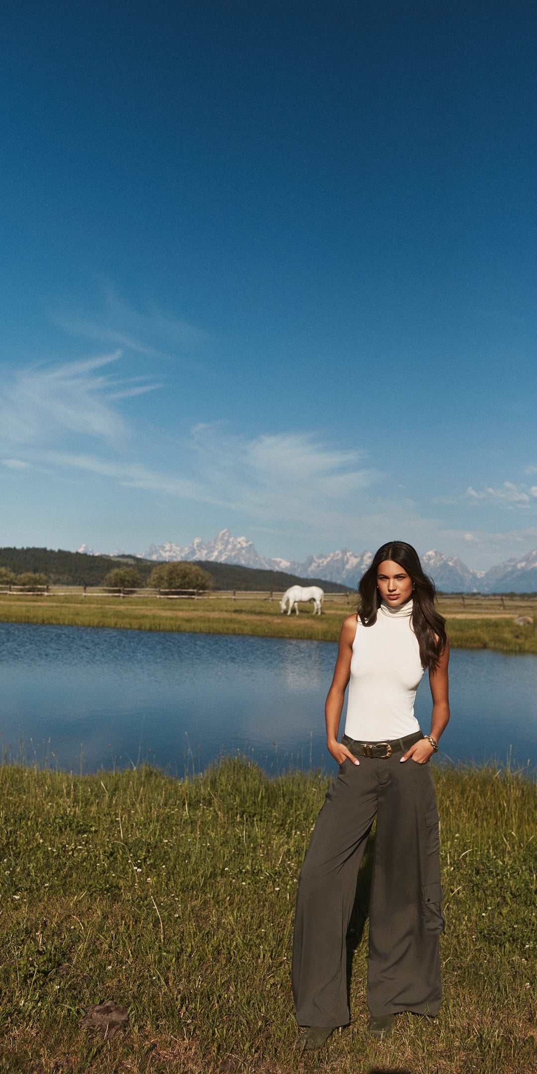 A woman in a white sleeveless top and wide-leg pants stands by a pond in a grassy field, with mountains and a white horse grazing peacefully in the background under a clear blue sky.