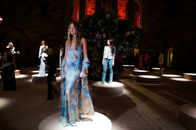 Several women stand on round platforms in a dimly lit room, showcasing stylish clothing. The central woman wears a flowing, pastel-colored dress. A large floral arrangement and spectators are visible in the background.