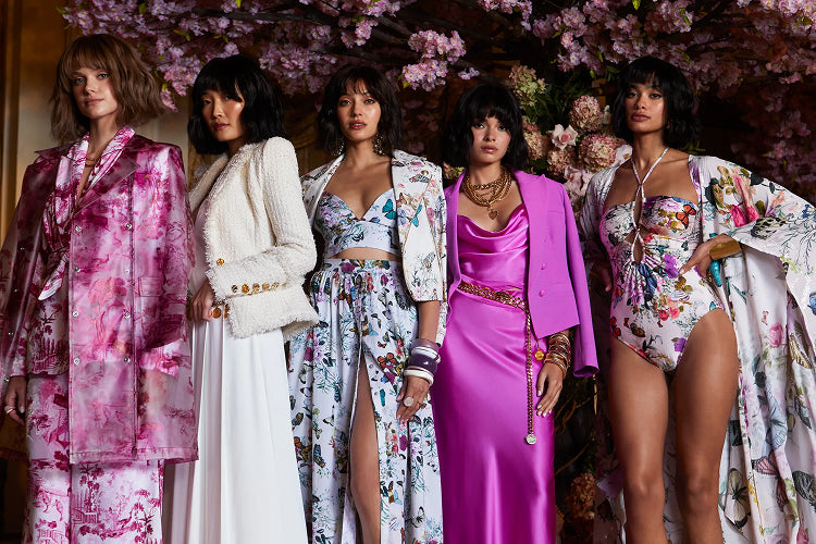 Five women stand in a row wearing vibrant, floral-patterned and pink outfits, including dresses, a suit, and a swimsuit, with a backdrop of blooming pink flowers.