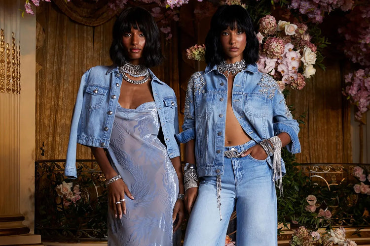 Two women with short dark hair pose in a lavish, flower-filled room. Both wear embellished denim jackets and silver jewelry; one pairs her jacket with a sheer dress, while the other wears jeans and a belt with a bare midriff.