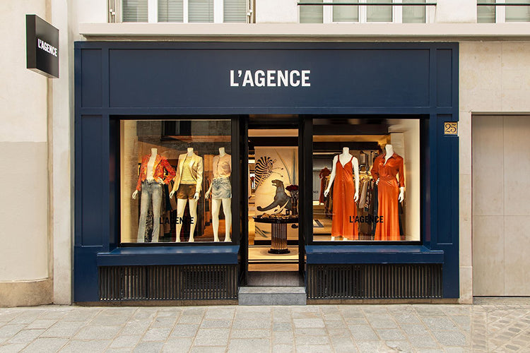 The storefront of L’AGENCE displays four mannequins dressed in stylish women’s clothing, including dresses and jackets, behind large windows with a navy blue sign above the entrance.