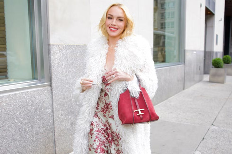 A woman with blonde hair wearing a white furry coat over a floral dress smiles while walking on a city sidewalk. She carries a red handbag and stands in front of a modern building.