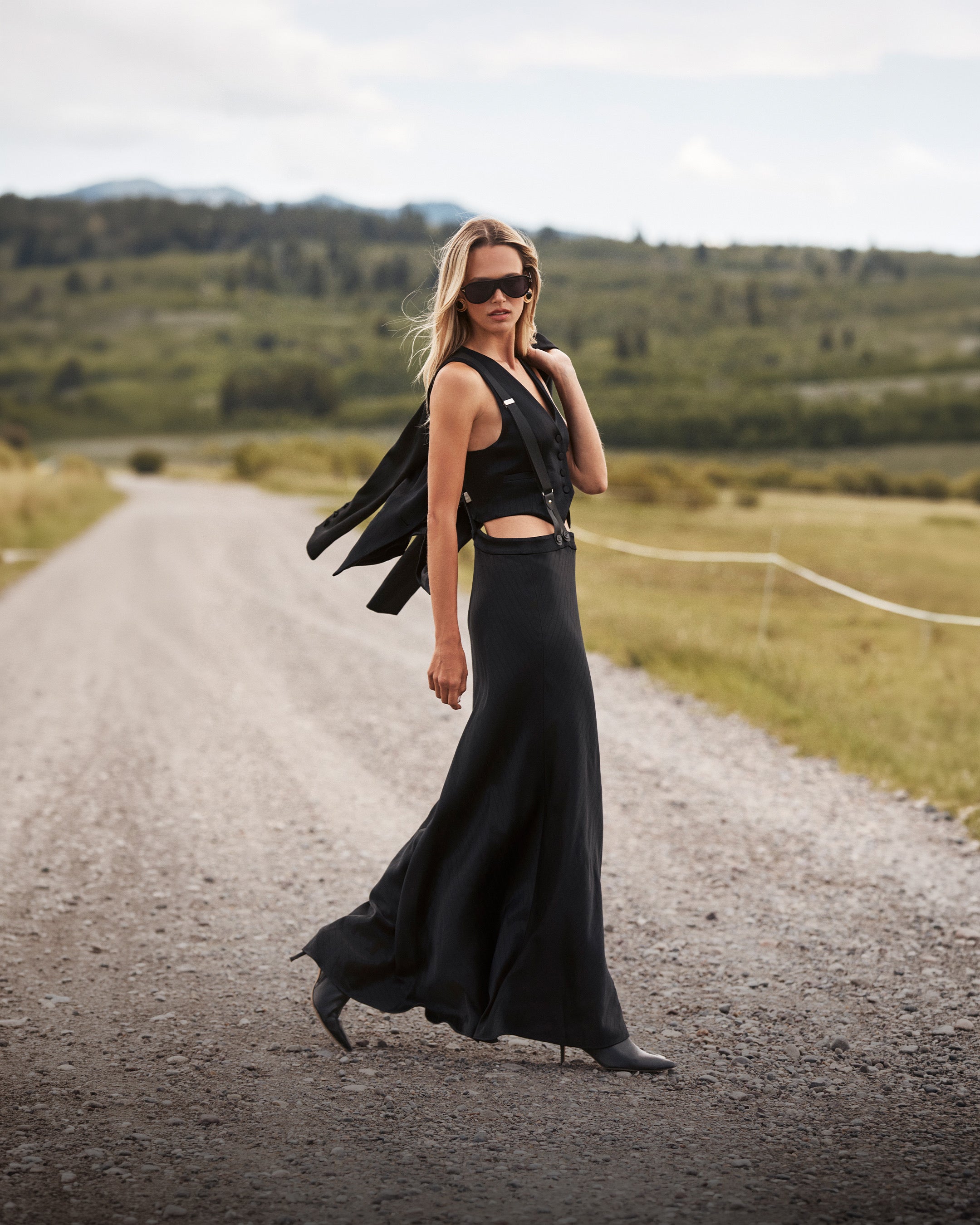 A woman in a long black dress and sunglasses walks on a gravel road through a rural landscape, with rolling green hills and cloudy skies stretching out in the background.