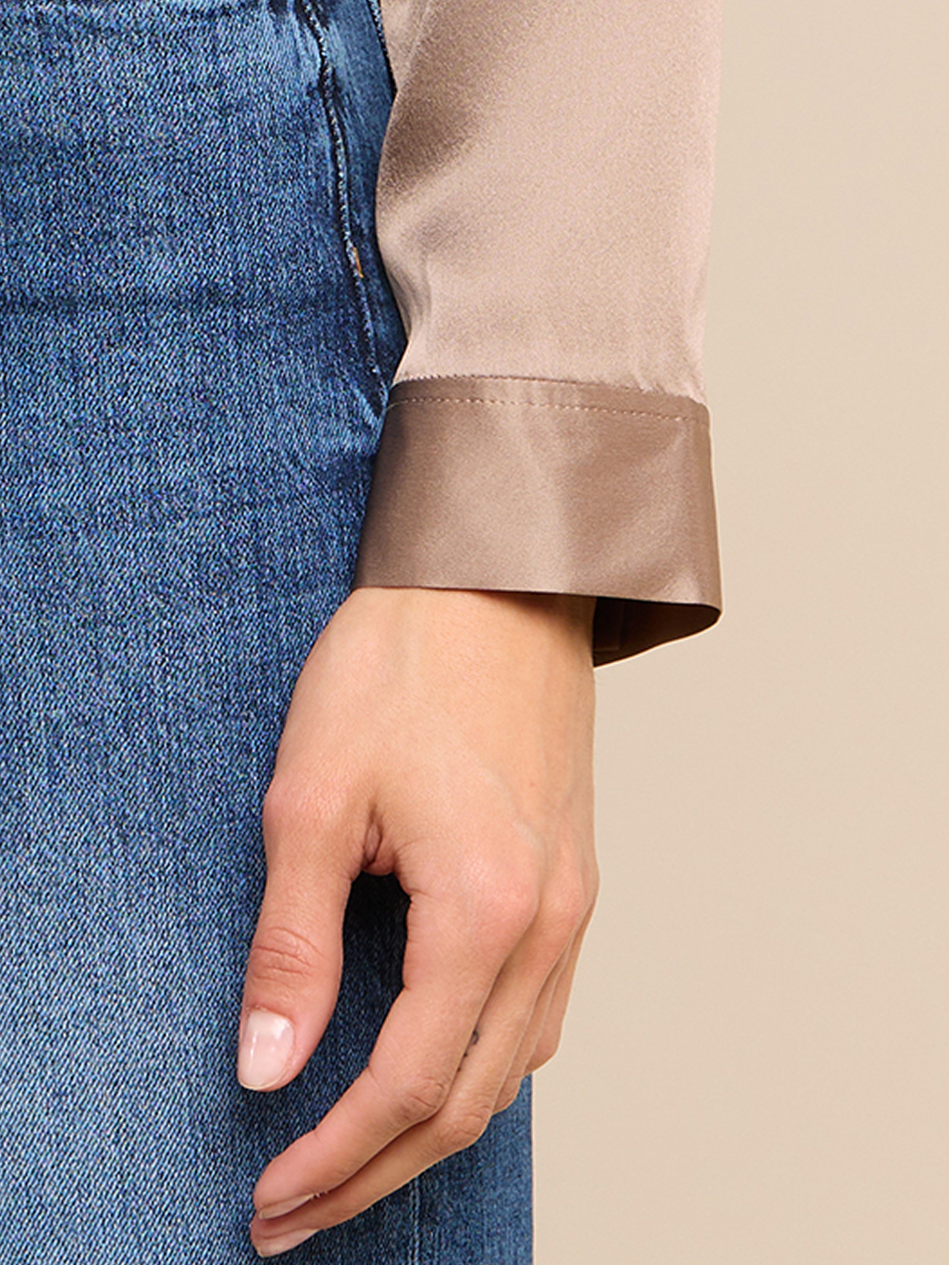 A close-up of a person’s left hand resting against their thigh, wearing a beige satin long-sleeve top and blue denim jeans.